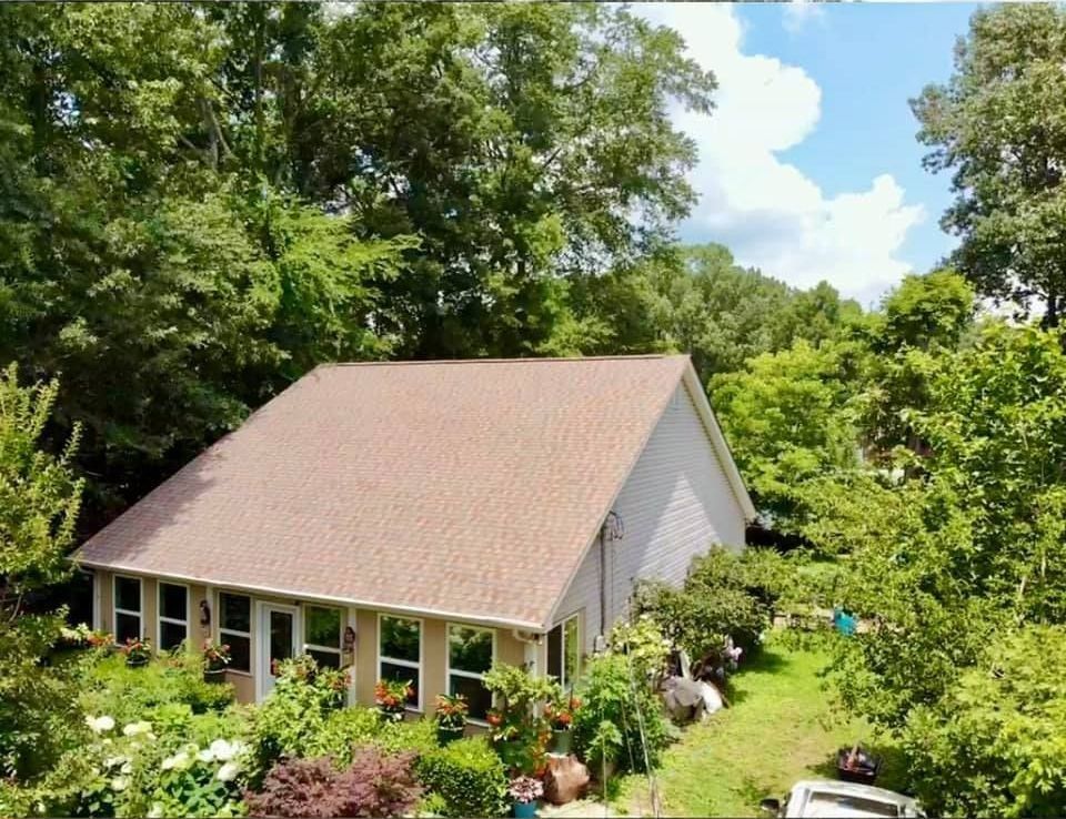 A light-colored house with a brown shingled roof surrounded by lush trees and a garden on a sunny day.