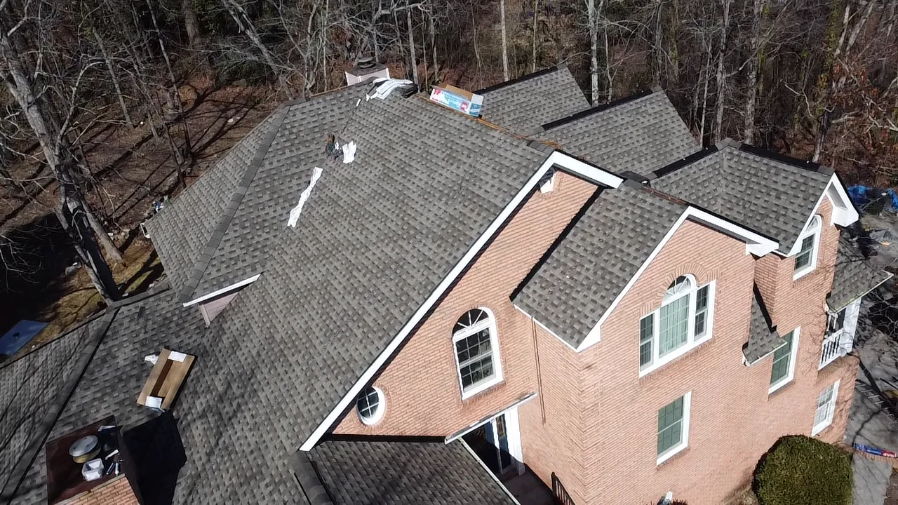 An aerial view of a multi-story brick house with a complex dark shingled roof surrounded by trees.
