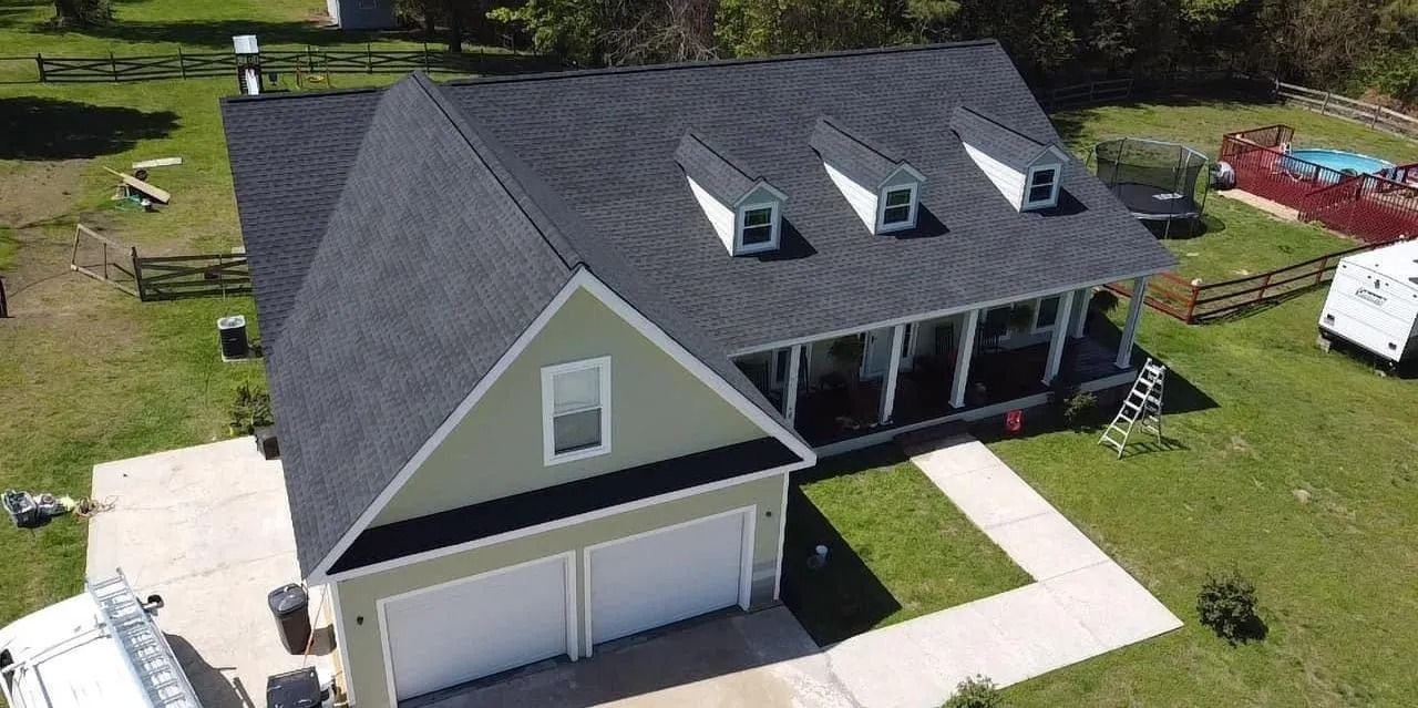 Aerial view of a light green house with a dark shingled roof, three dormers, and a two-car garage on a grassy lot.