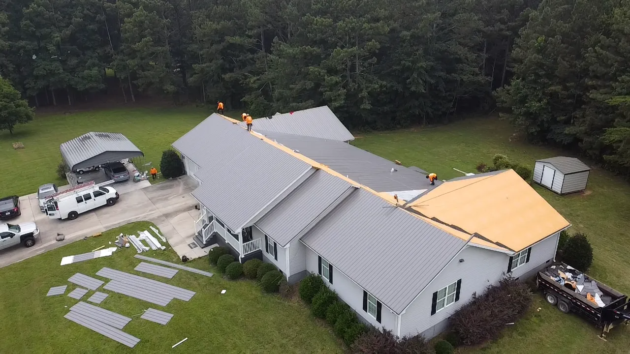 An aerial view of workers installing a metal roof on a residential house surrounded by trees, with panels laid on the lawn.