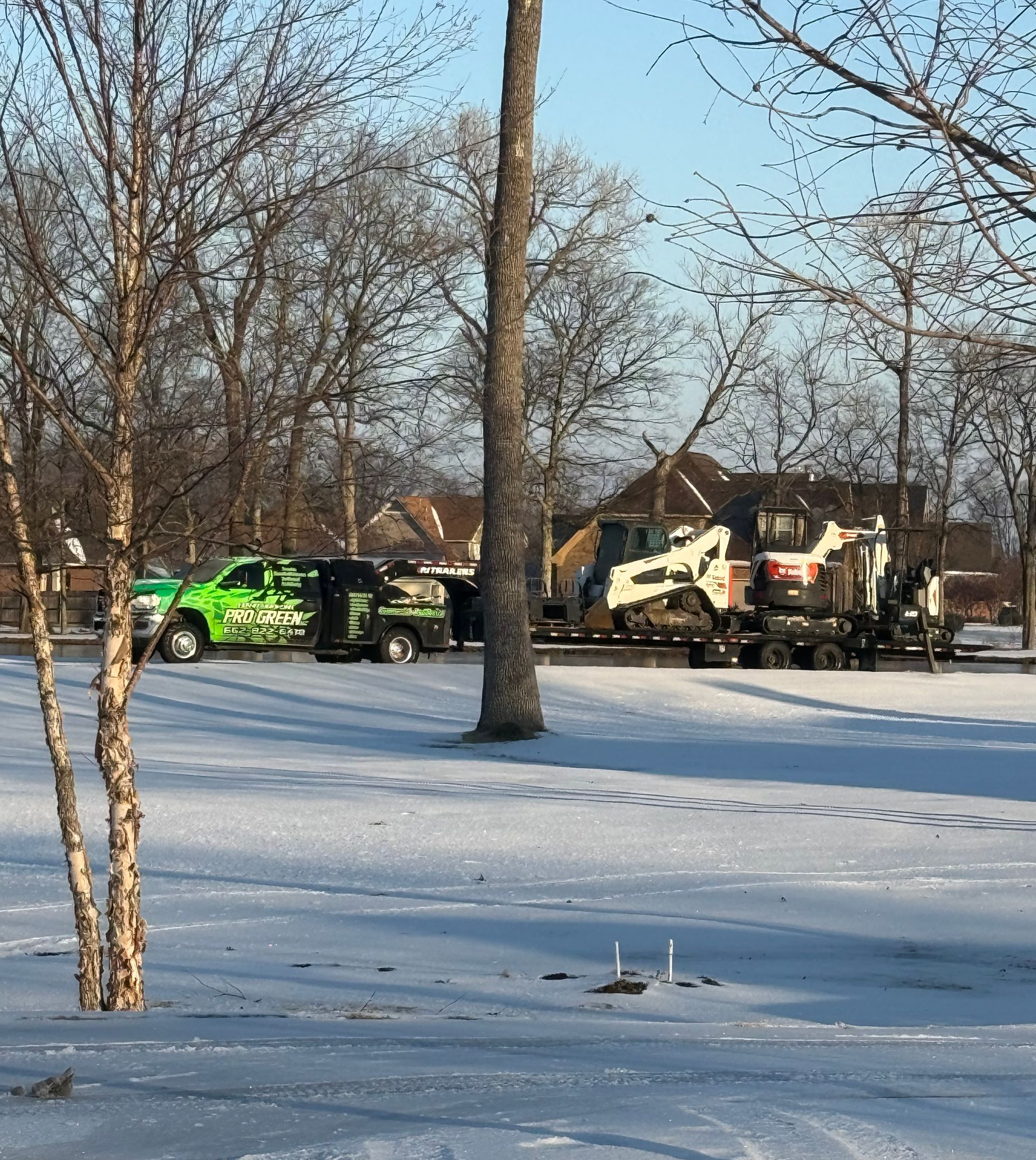 Snowy scene with green truck, trailer carrying equipment, trees, and house.