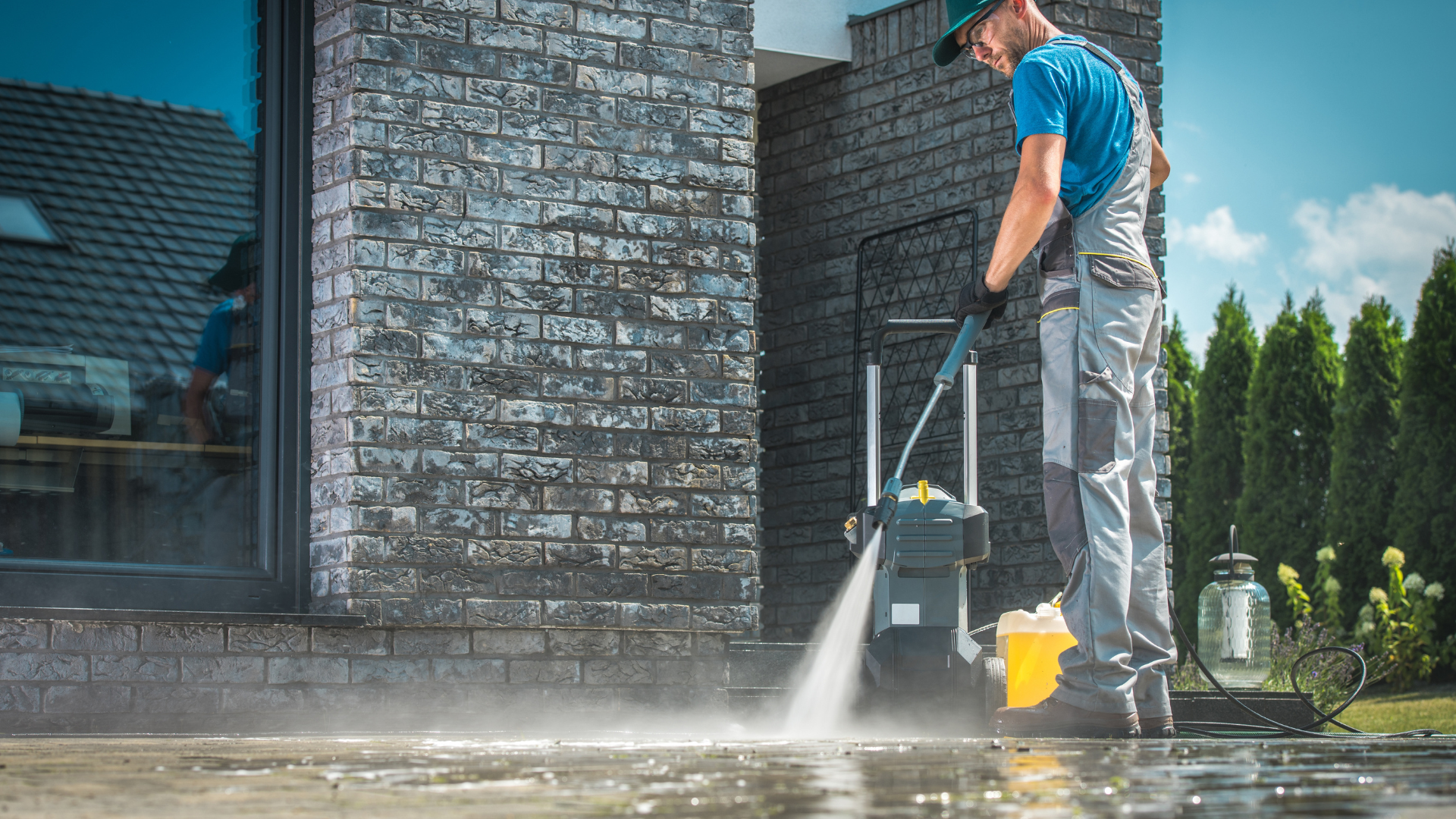 A person in work coveralls uses a high-pressure washer to clean a paved patio in front of a modern brick house.
