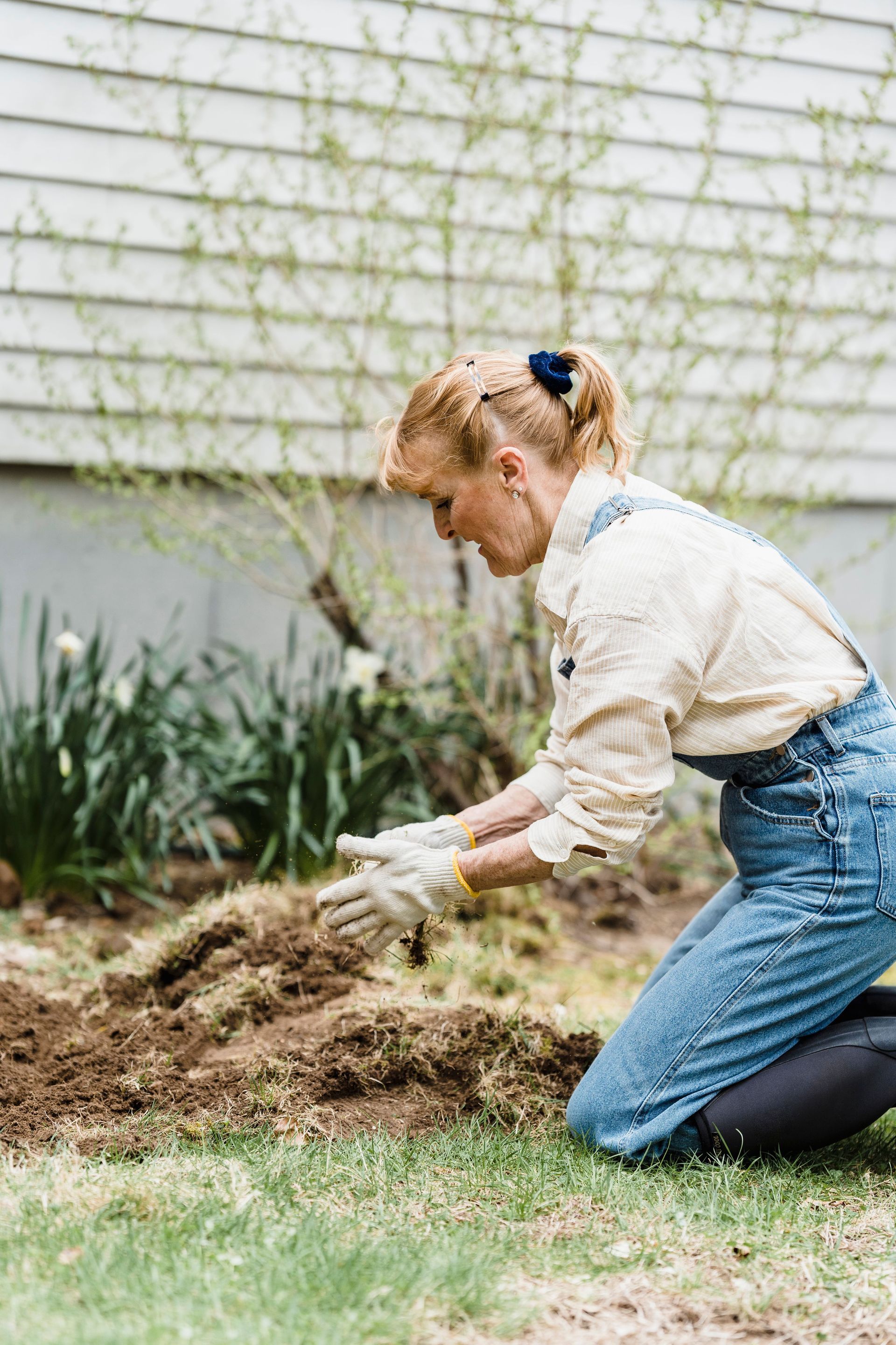 Person kneeling in a garden, pulling up weeds beside a house with a rake nearby.