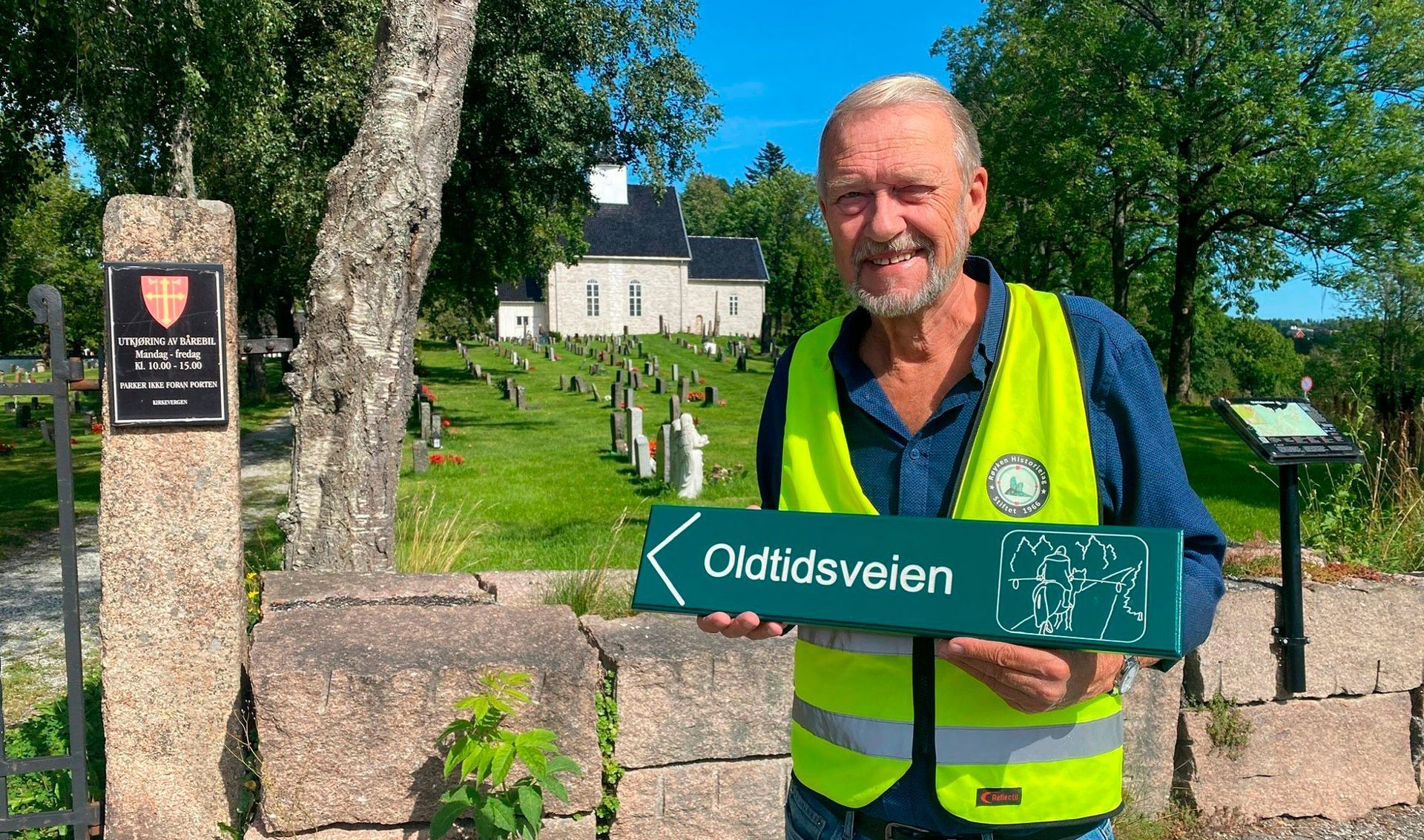 Mann som holder et skilt med teksten «Oldtidsveien» foran en kirke, iført sikkerhetsvest.