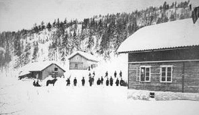 Vinterscene med snødekte bygninger og en gruppe mennesker samlet foran. Trær kantar åsen i bakgrunnen.