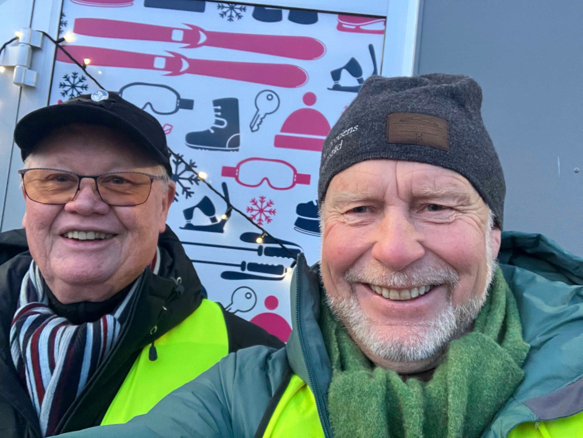 To smilende menn poserer foran et skilt med vintersporttema, iført luer, skjerf og frakker.