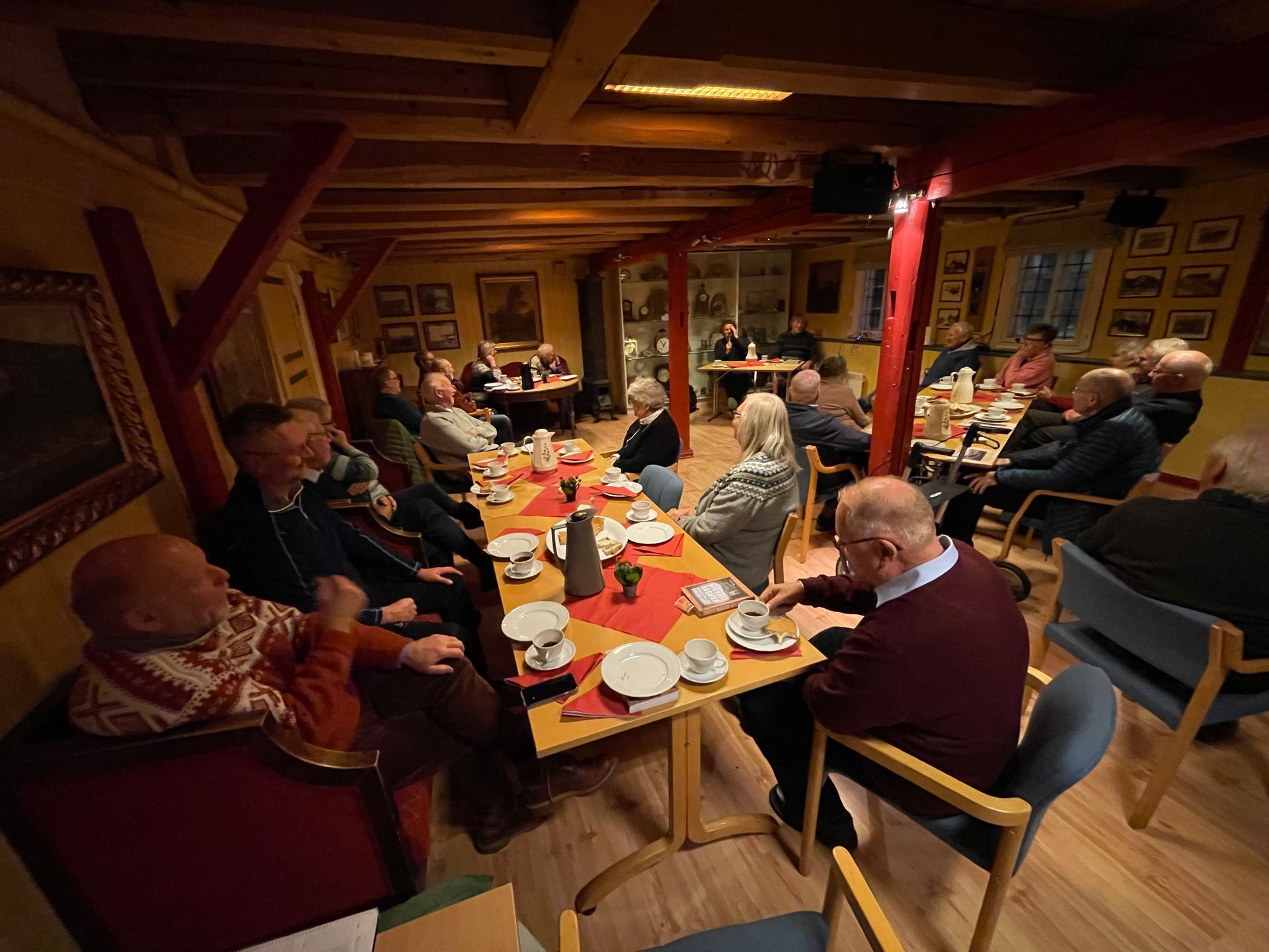 Folk sitter ved bord i et rom og lytter til en presentasjon. Trebjelker og kunst på veggene.