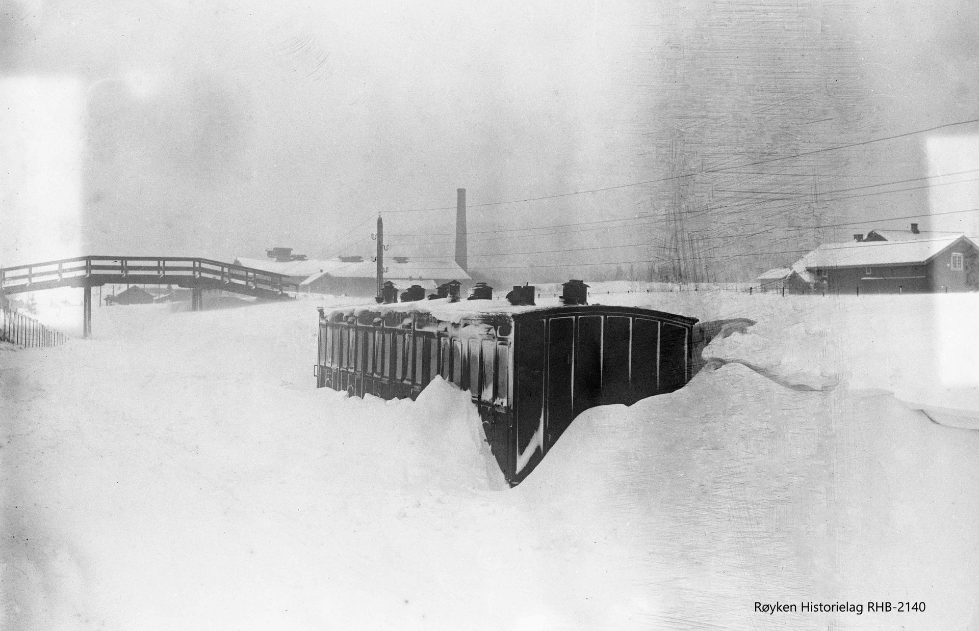 Togvogner begravd i dyp snø, bro, bygninger i bakgrunnen. Dystert vinterlandskap.