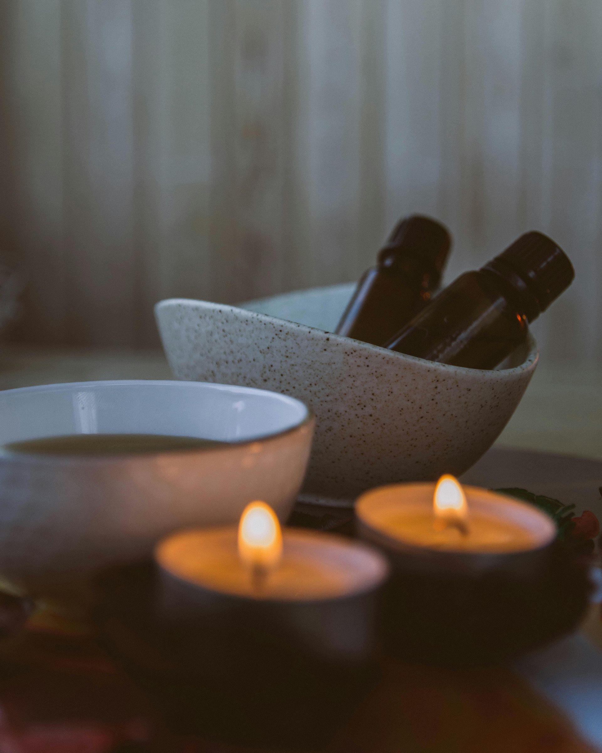 Two lit candles near bowls, one with oil, others holding dark bottles, wood background.