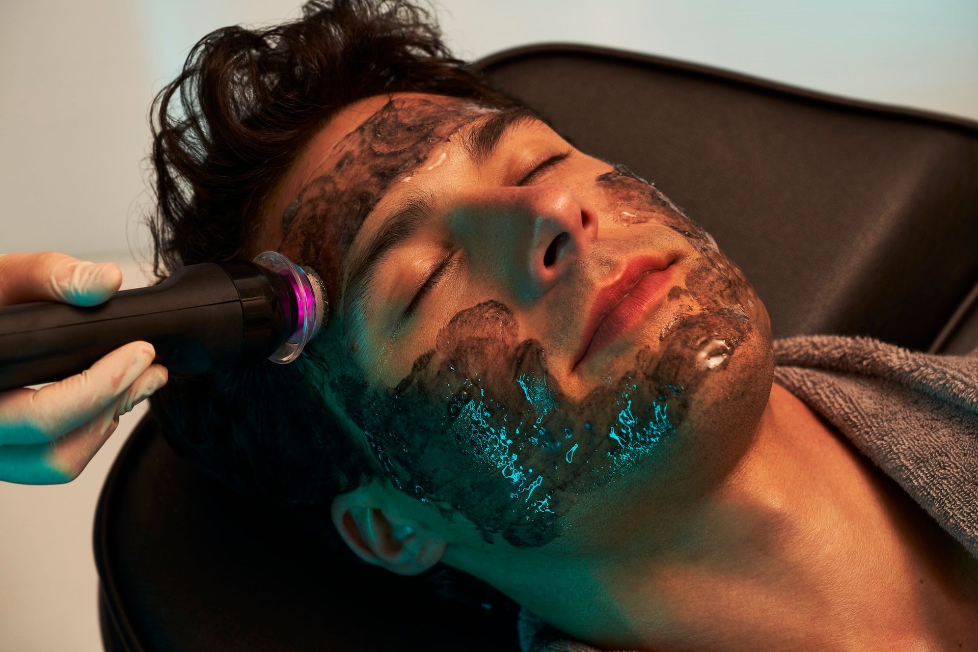 Man undergoing a facial treatment with a black mask. Light shone on his face. Person in a chair.