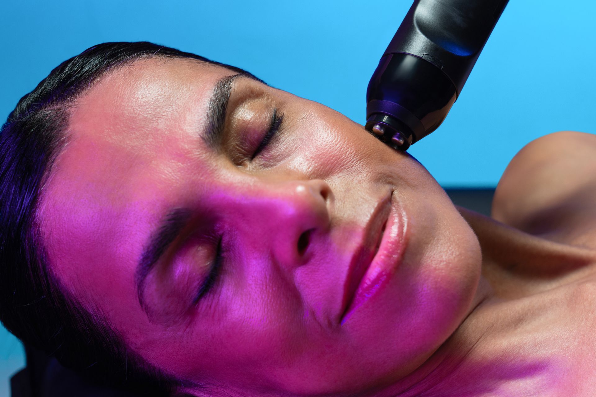 Woman receiving facial treatment with device; pink light, blue background.