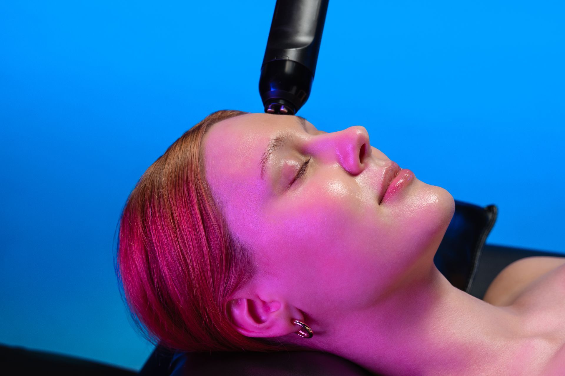 Woman receiving facial treatment with device, eyes closed, pink light, blue background.