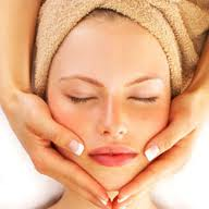 Woman with closed eyes receiving facial massage, hands framing face, wrapped in towel.