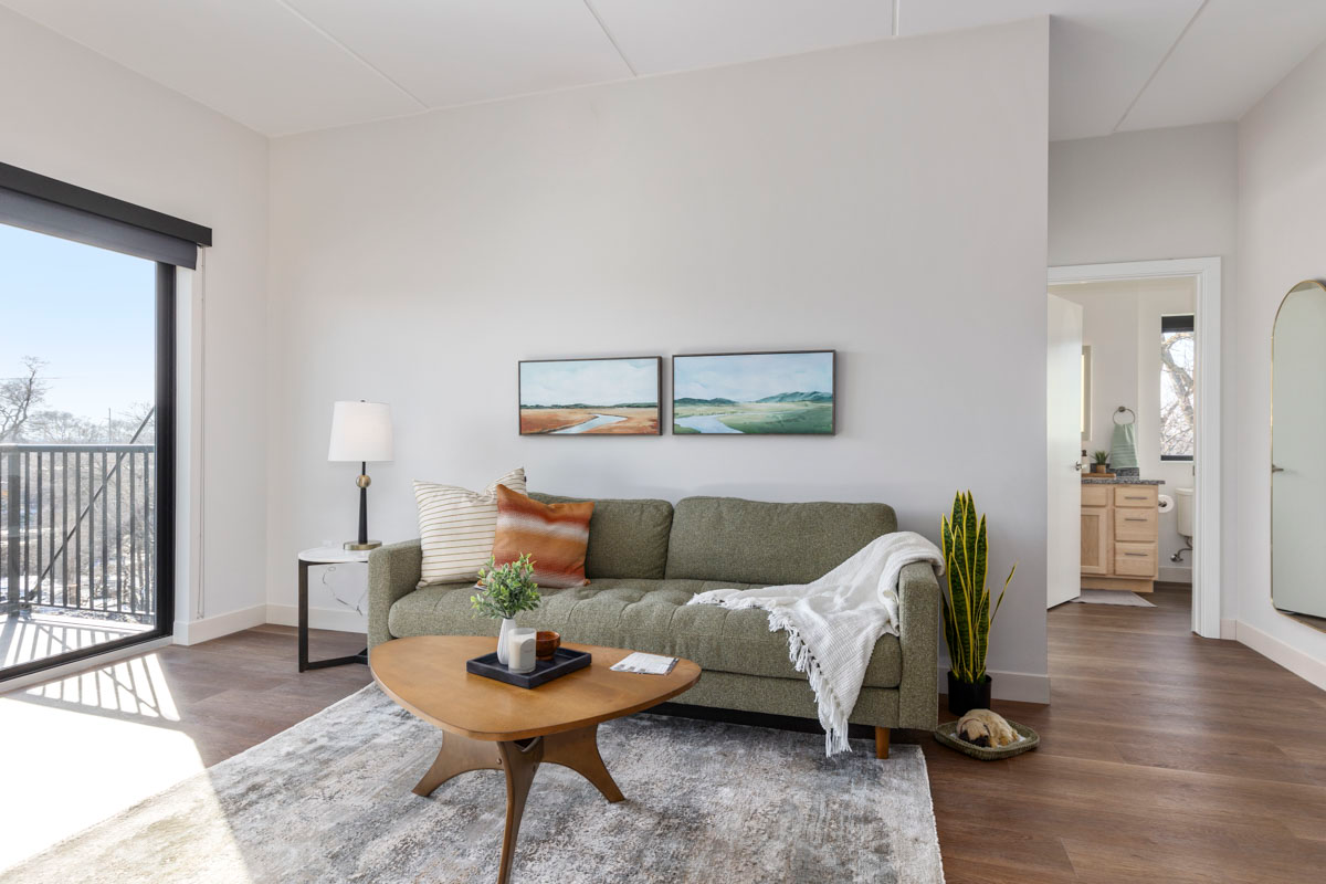 Bright living room with green sofa, round wood table, rug, floor lamp, and sliding glass door to balcony