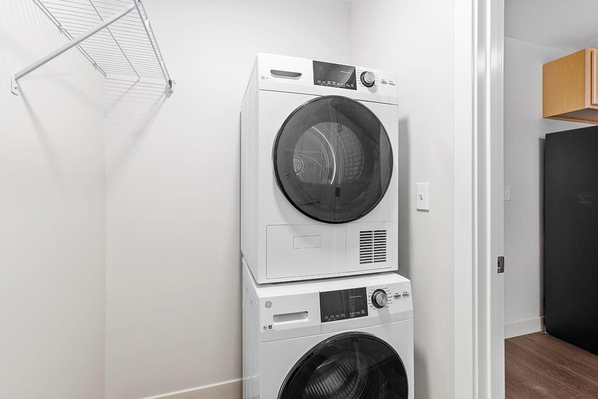 Stacked white washer and dryer in a small laundry closet with an ironing board on the left.
