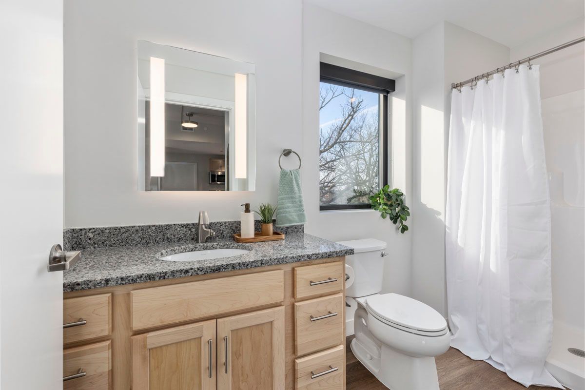 Bright bathroom with wood vanity, illuminated mirror, window, and shower curtain