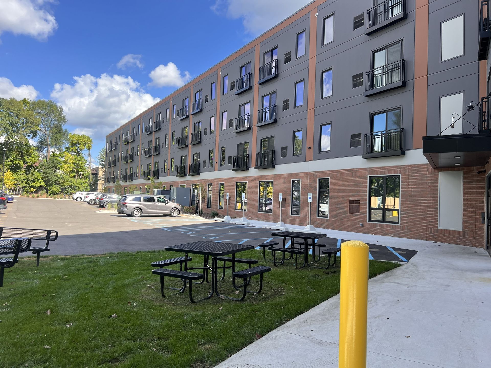 Apartment building with outdoor picnic tables, grassy courtyard, and parked cars under a blue sky.