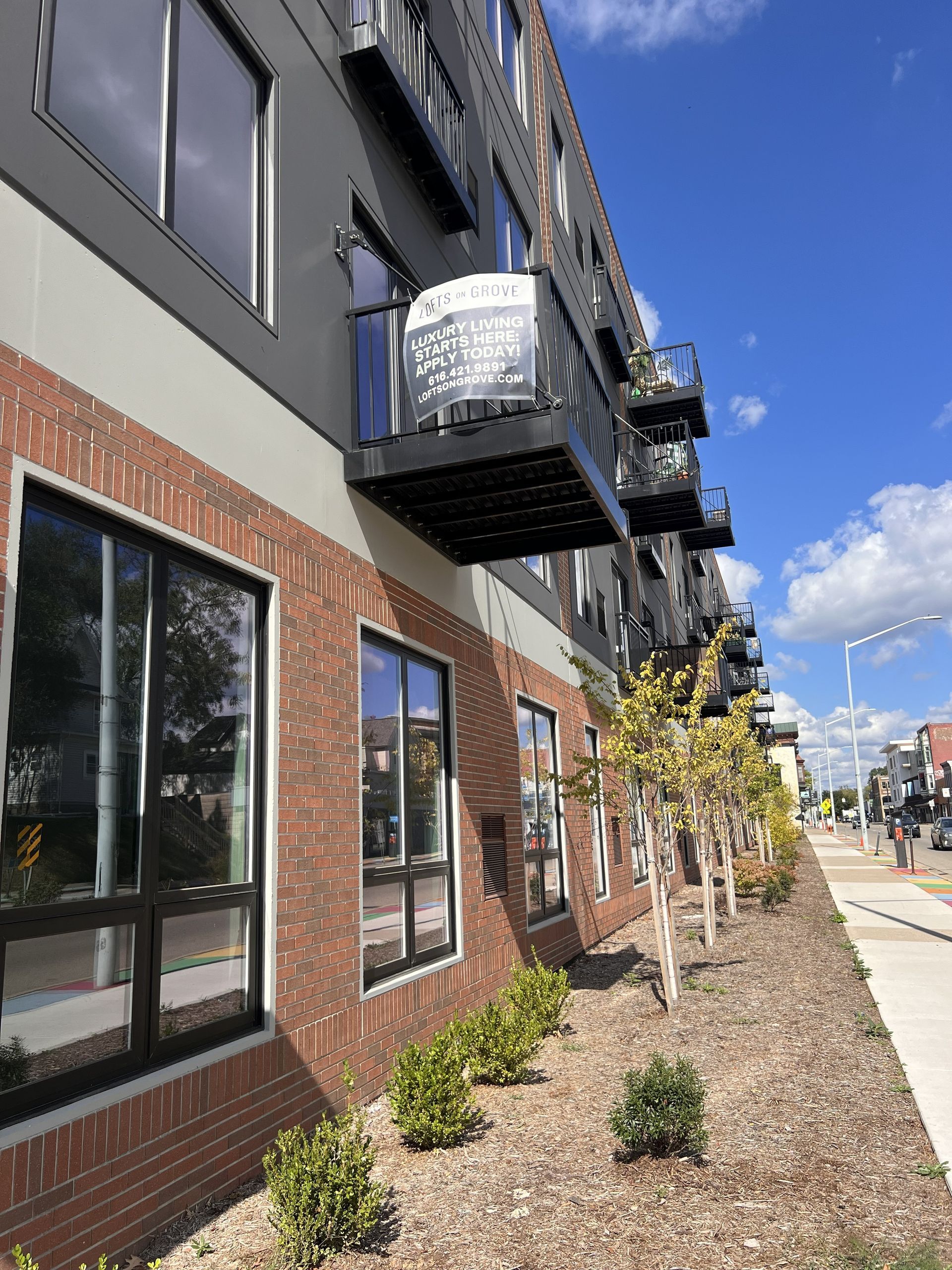 Modern apartment building with balconies, brick and gray facade, and a landscaped sidewalk under a blue sky.