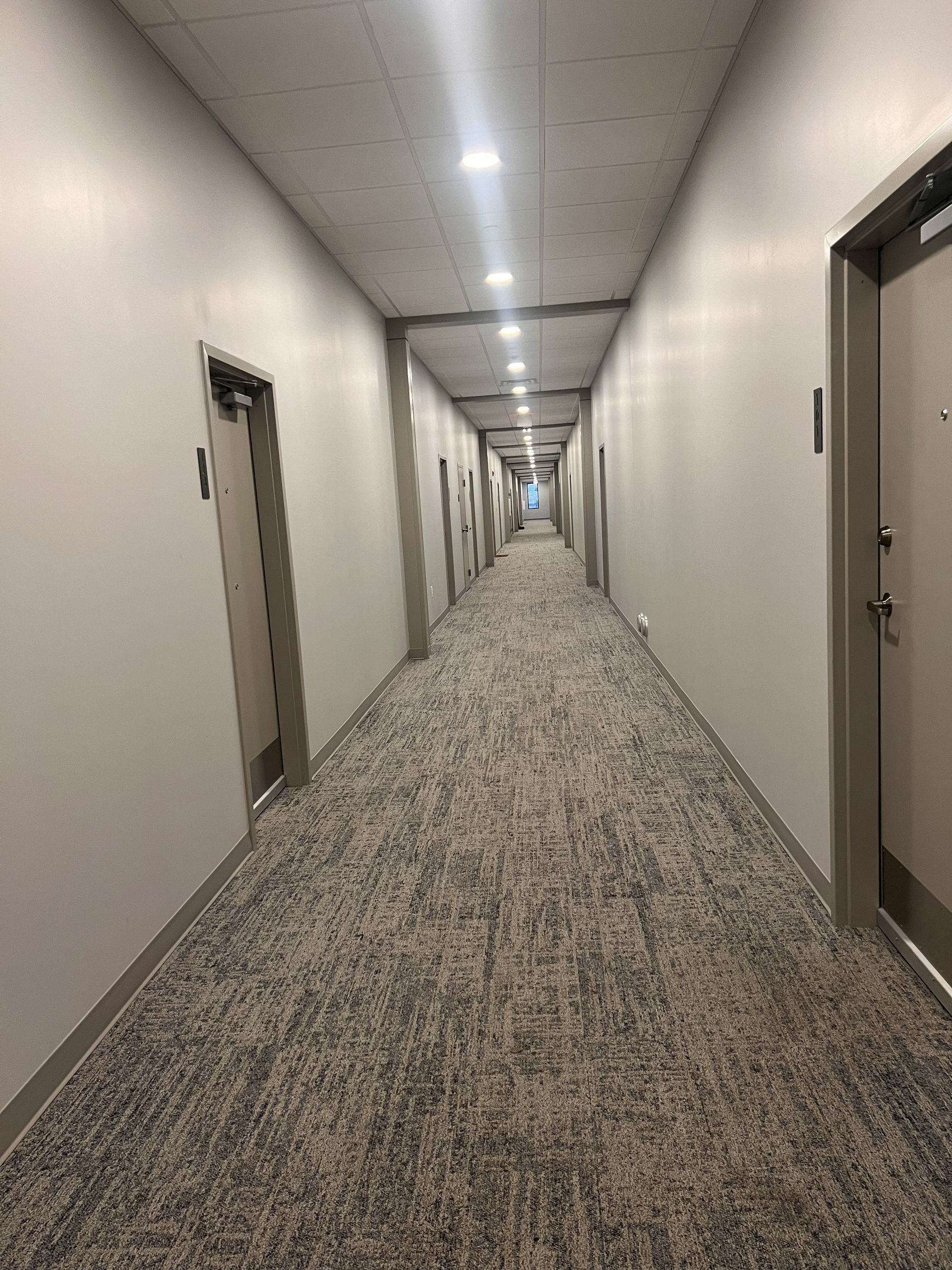 Long hotel-like hallway with beige walls, patterned carpet, and doors on both sides.