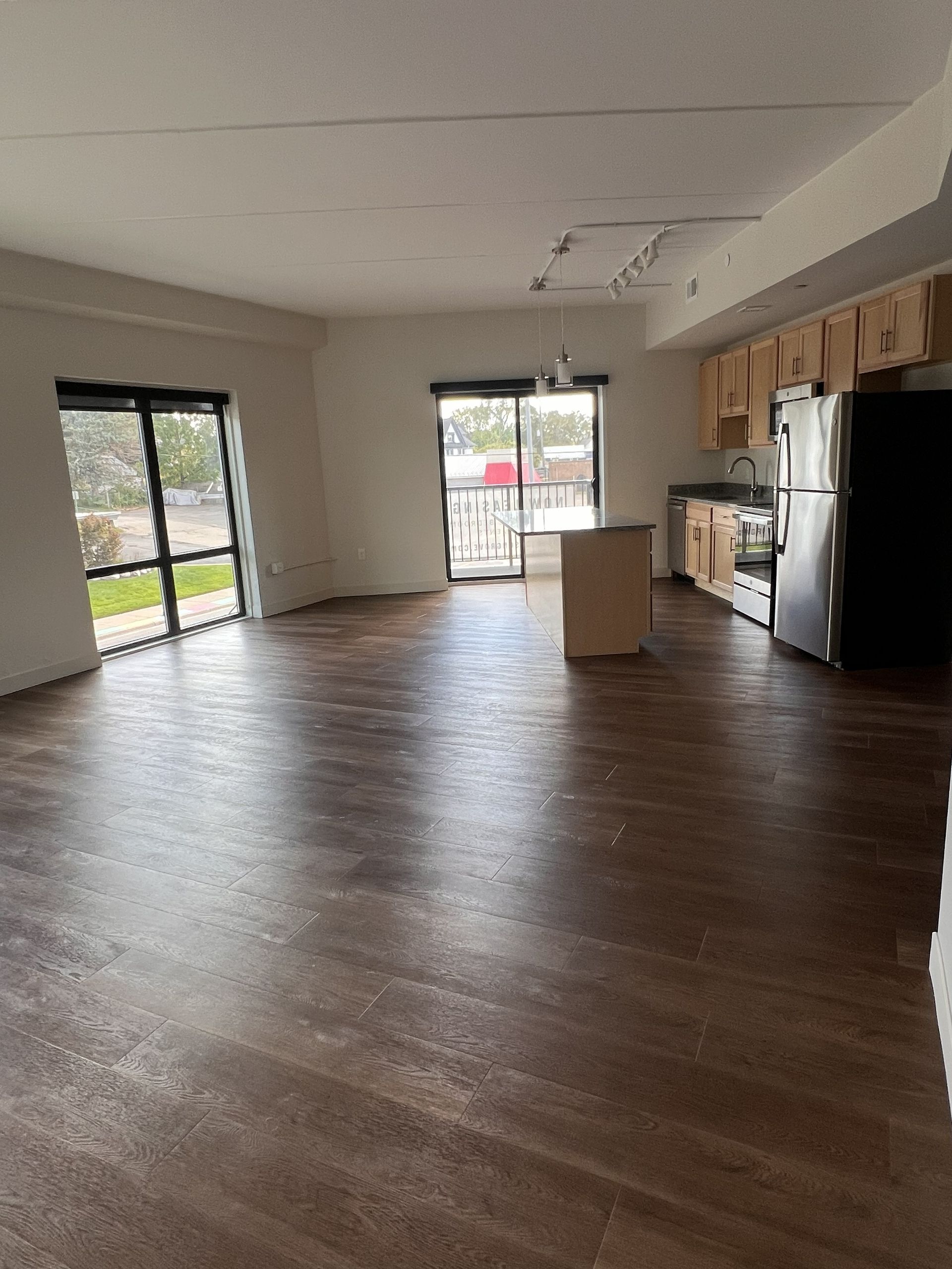 Empty open-plan kitchen and living area with wood floors, stainless fridge, and large sliding glass doors.