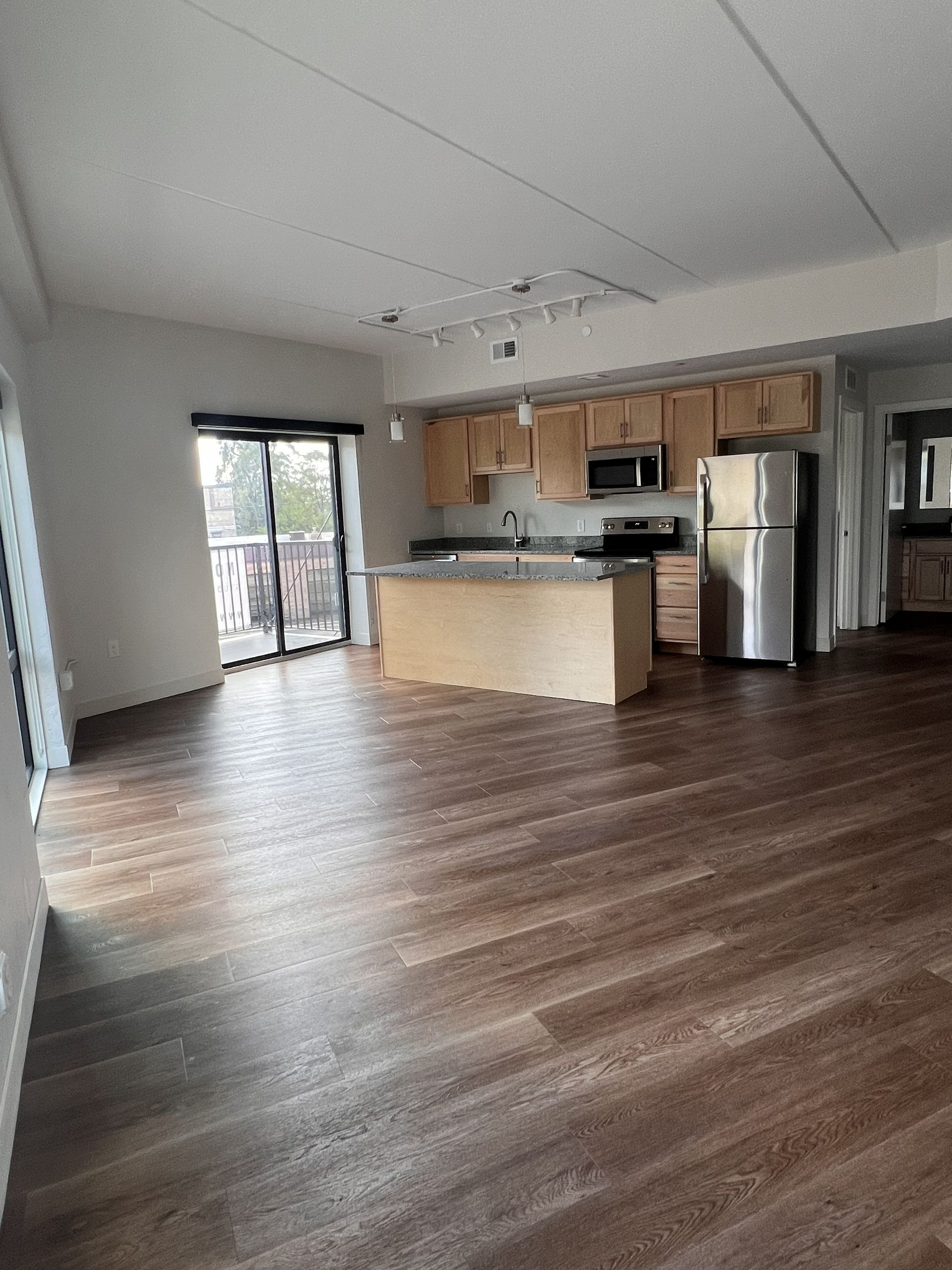 Empty modern kitchen and living area with wood floors, island, and stainless steel appliances