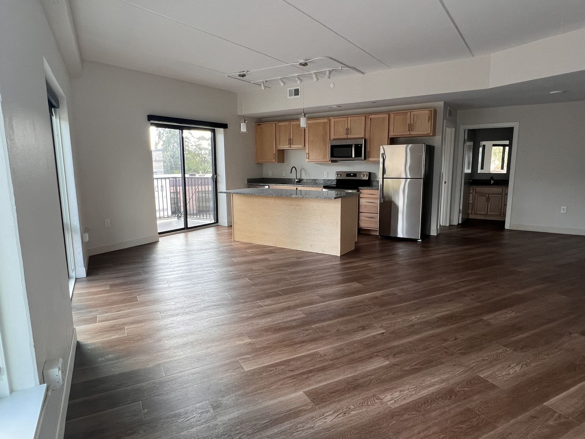 Empty open-plan living room and kitchen with wood floors, stainless fridge, and sliding glass door to balcony.
