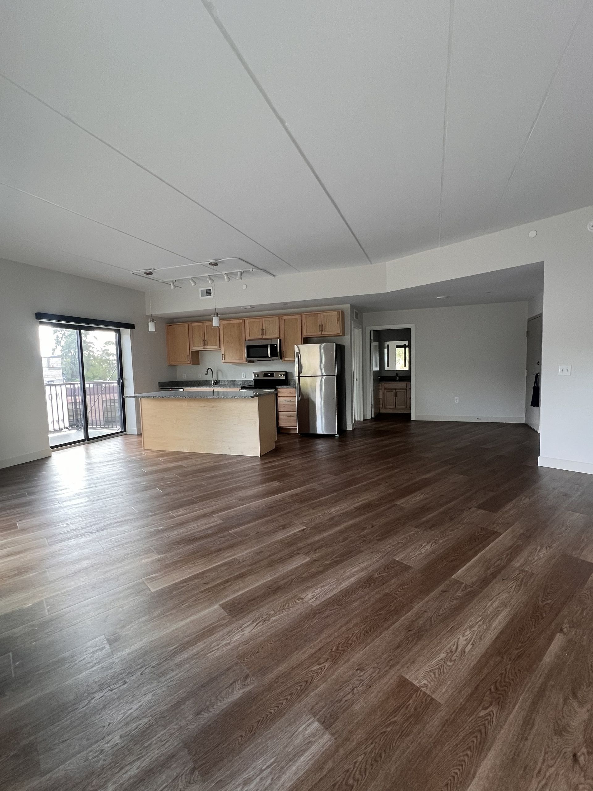 Empty open-plan living room with kitchen, wood floors, white walls, and sliding glass doors.