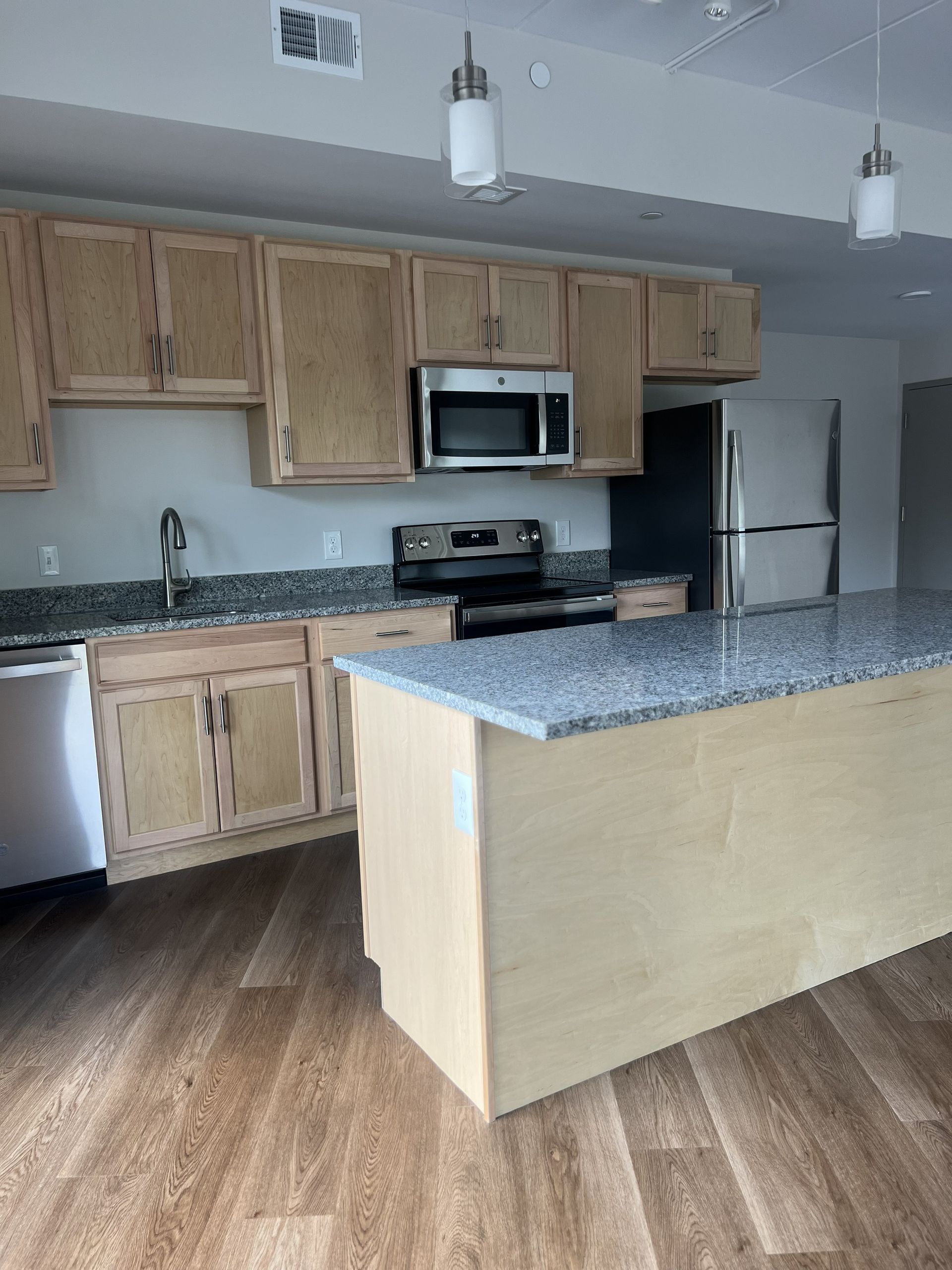 Empty kitchen with light wood cabinets, granite island, stainless appliances, and wood-look flooring.