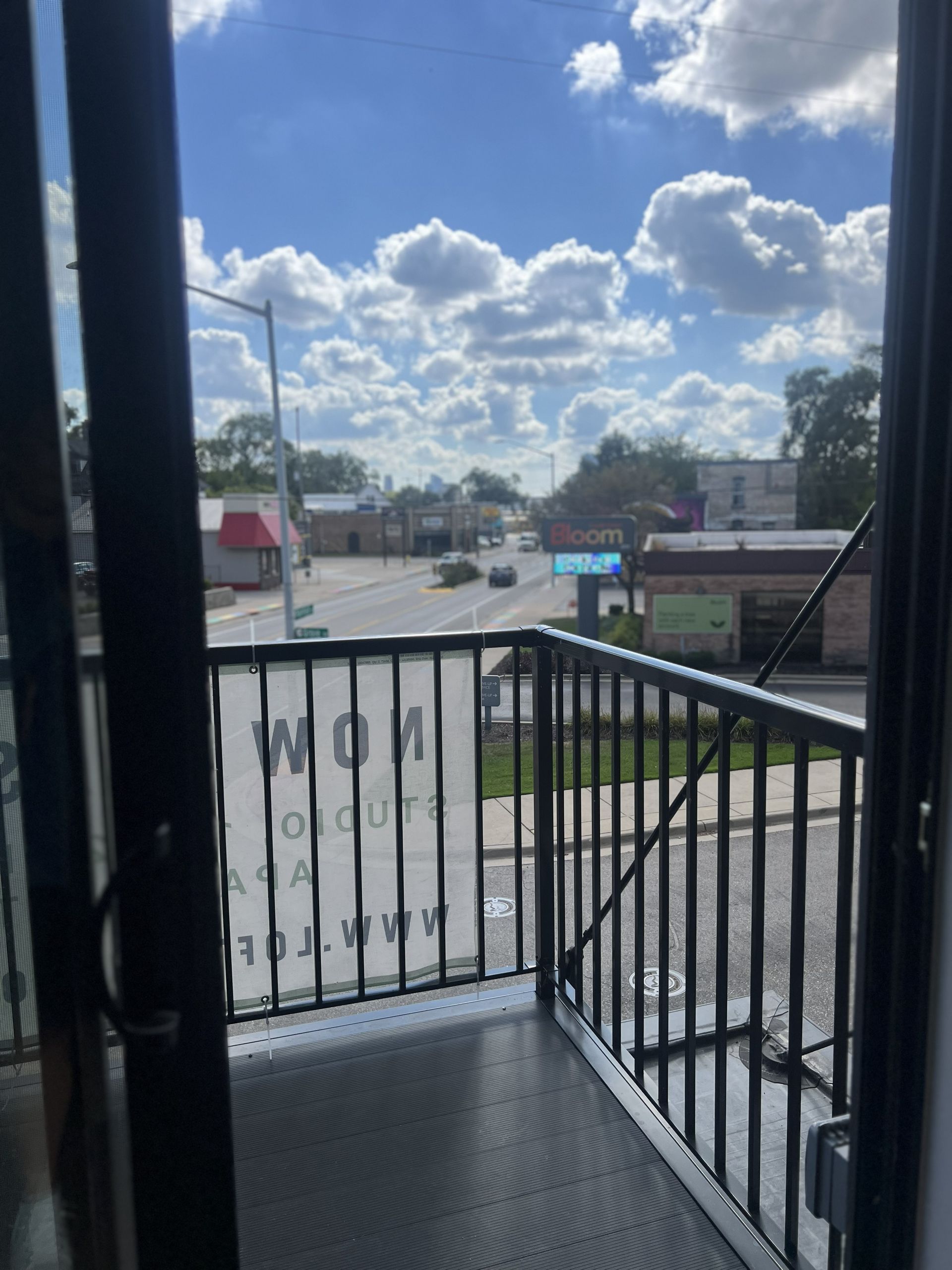 View from a balcony overlooking a street, railing in foreground, bright cloudy sky above.