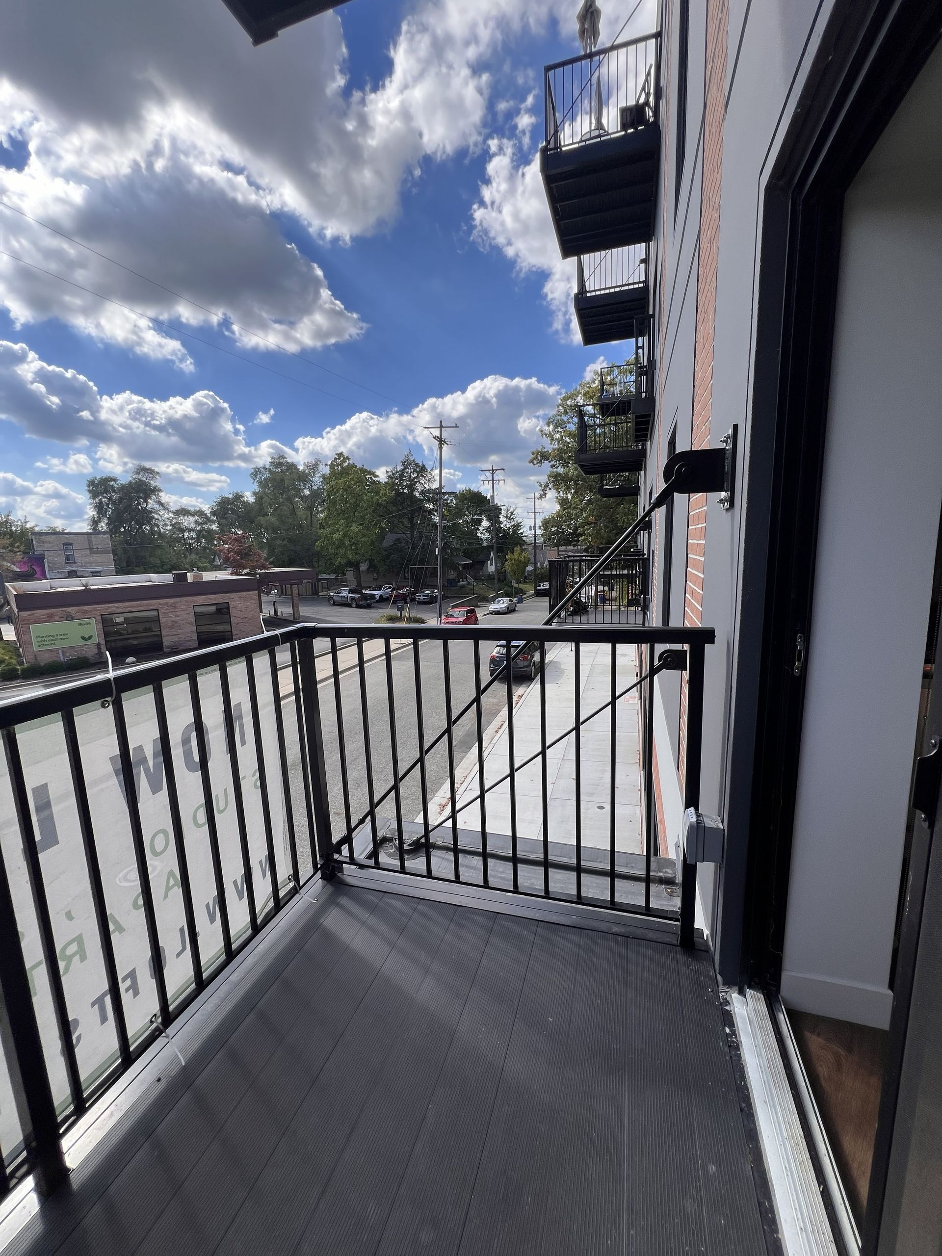 Small balcony with black railing overlooking a sunny street and apartment building.