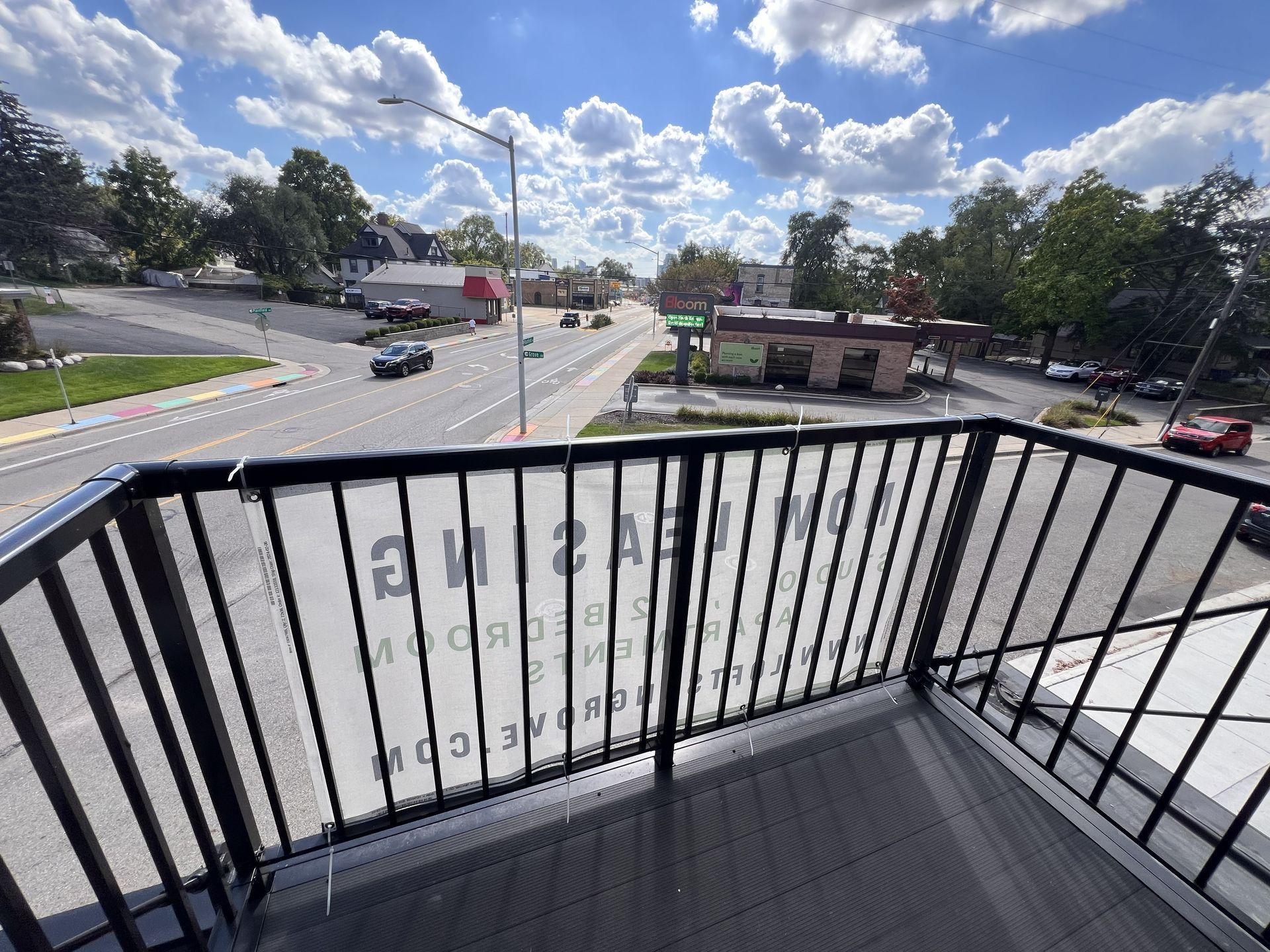 Balcony view of a sunny street intersection with cars, trees, and buildings under a blue sky.