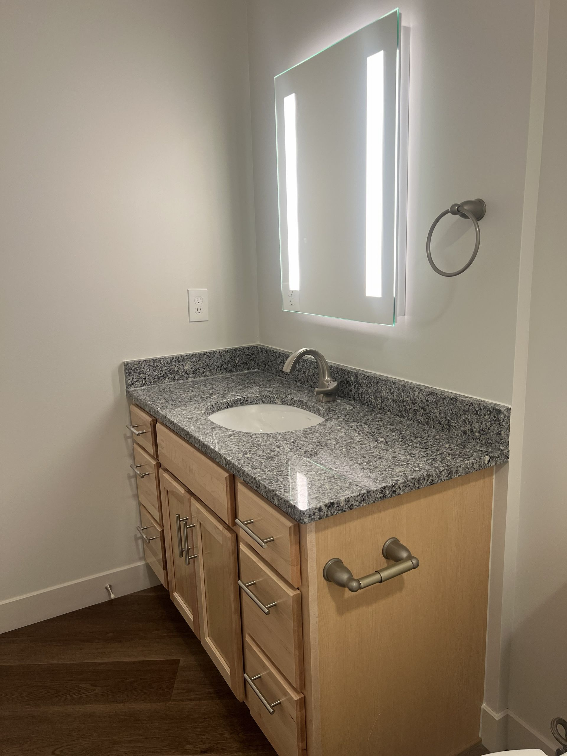 Bathroom vanity with speckled granite countertop, wood cabinets, oval sink, and lit mirror on a beige wall