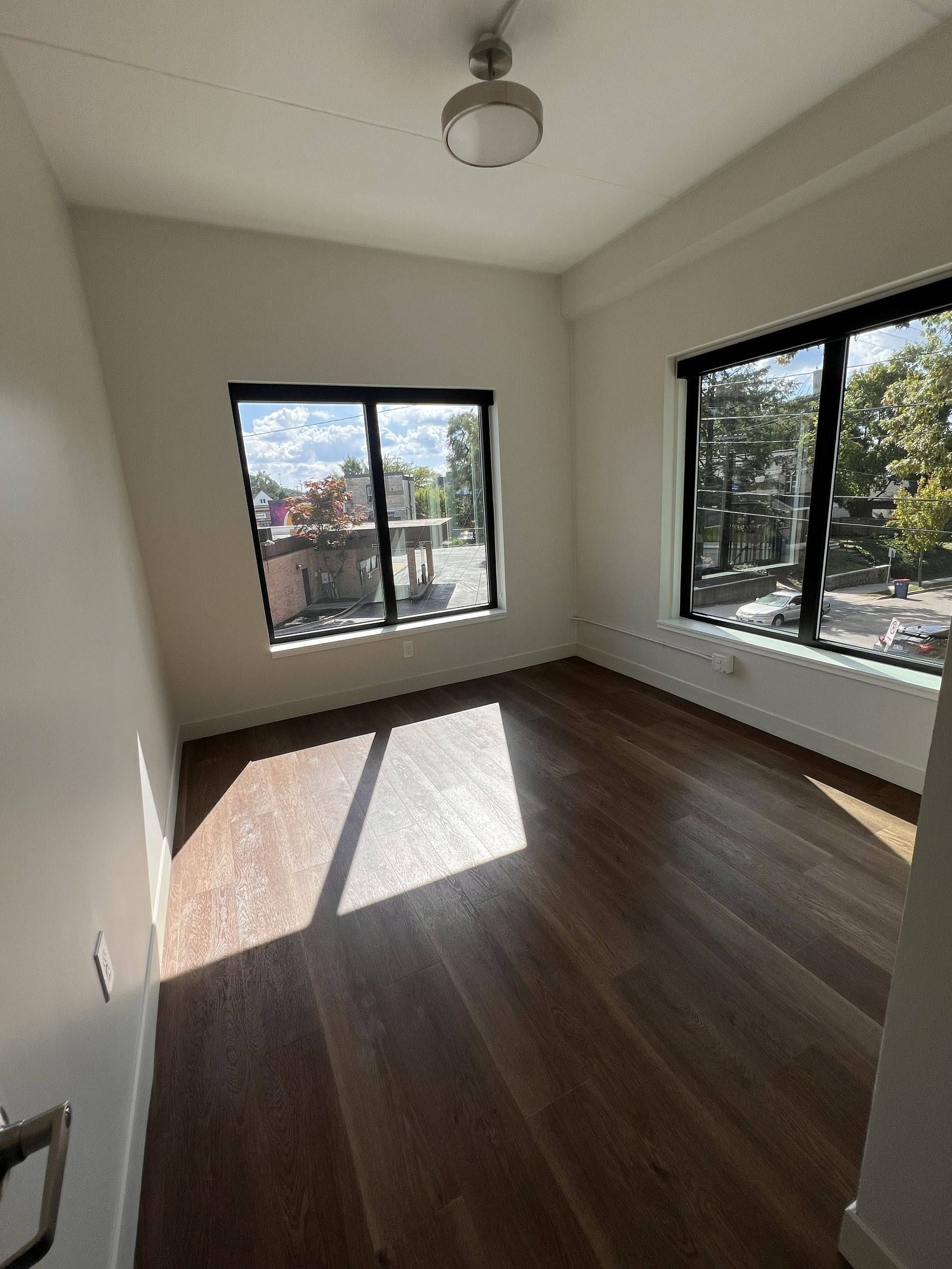 Empty sunlit room with dark wood floors and two large windows overlooking trees.