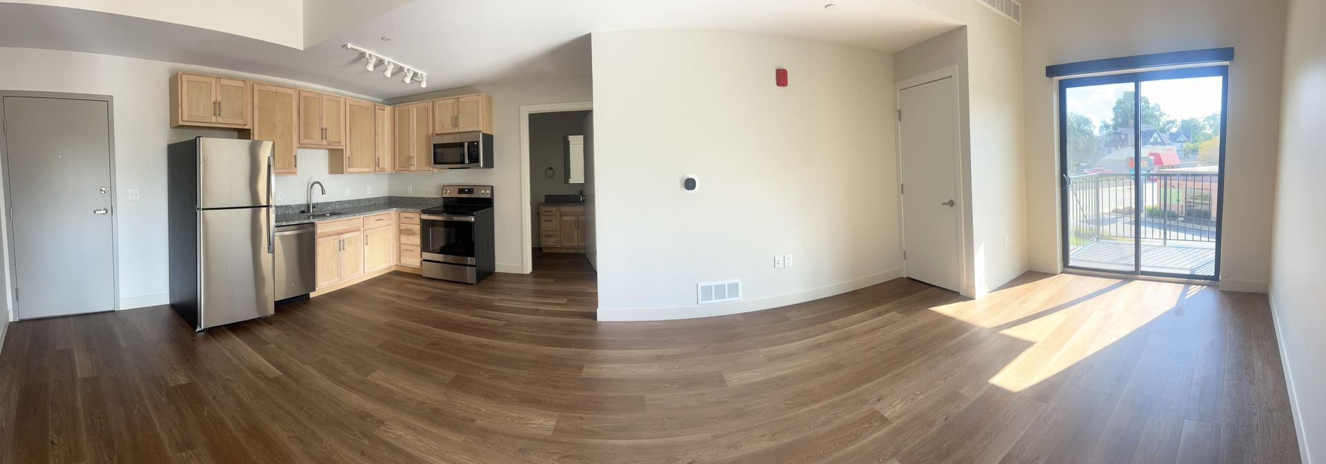 Empty modern apartment interior with kitchen, wood floors, and sliding glass door to balcony