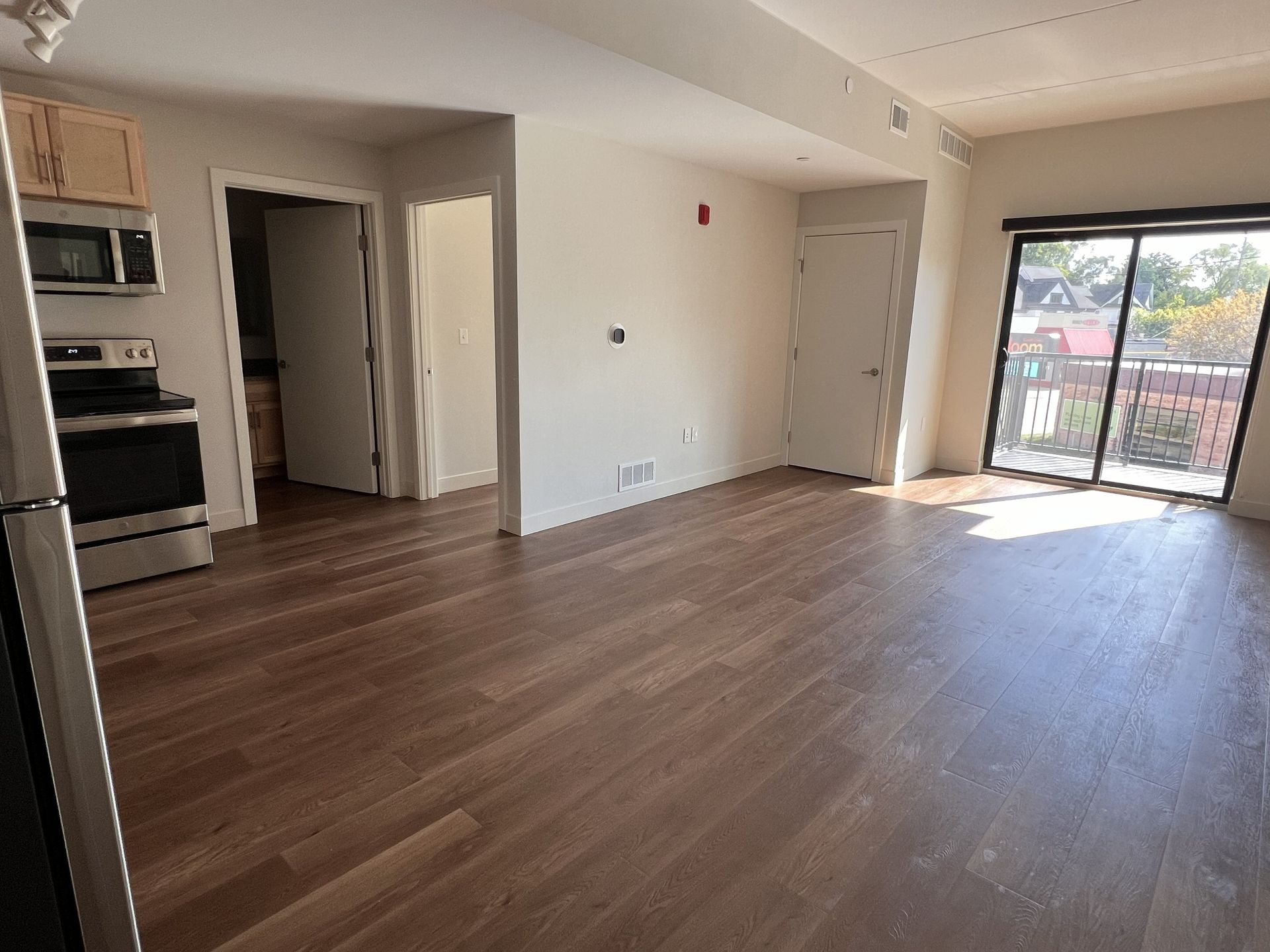 Empty modern living room with wood floors, white walls, kitchen nook, and sliding glass doors to a balcony.