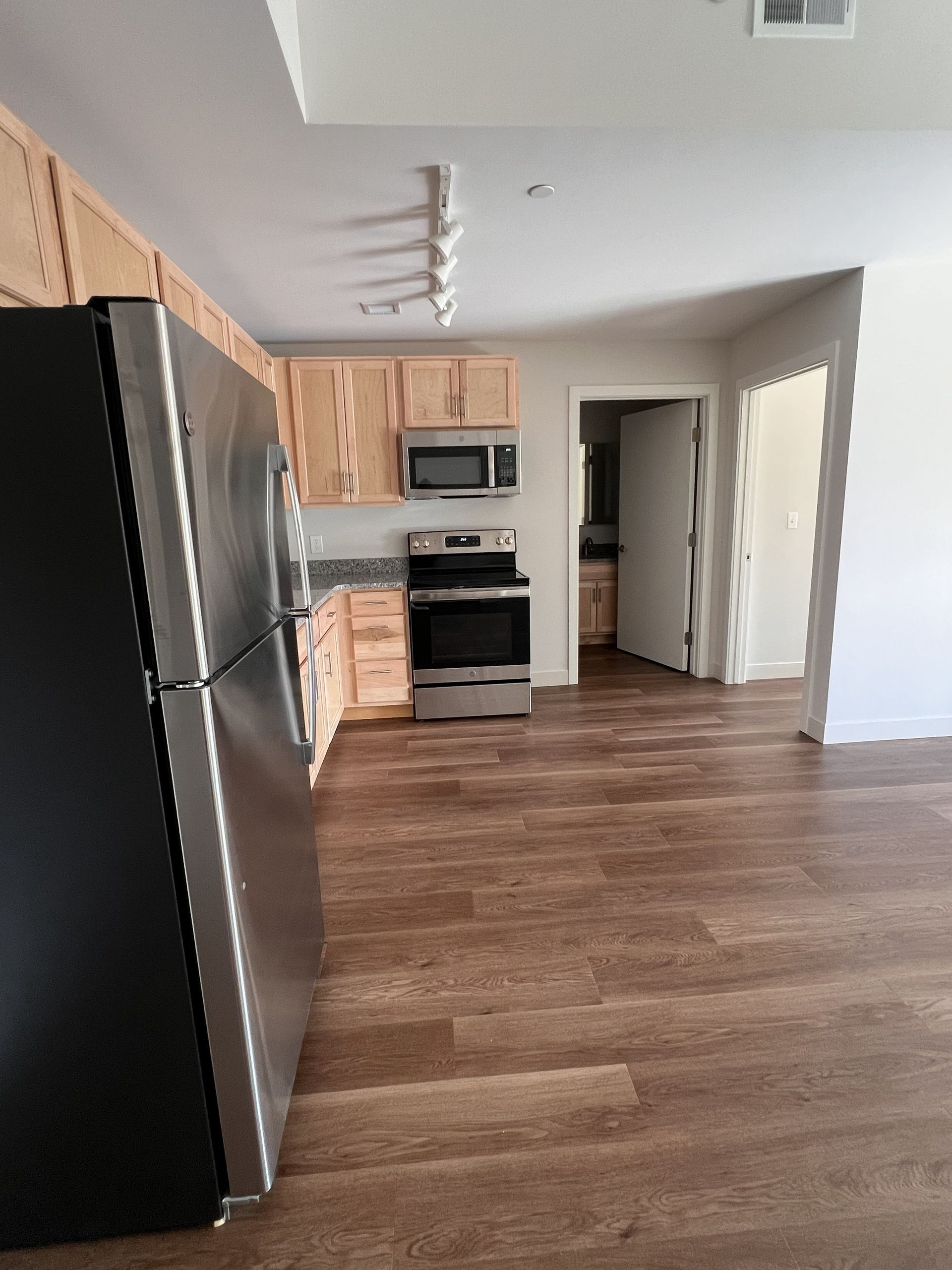 Empty kitchen with black refrigerator, light wood cabinets, white appliances, and wood-look flooring.