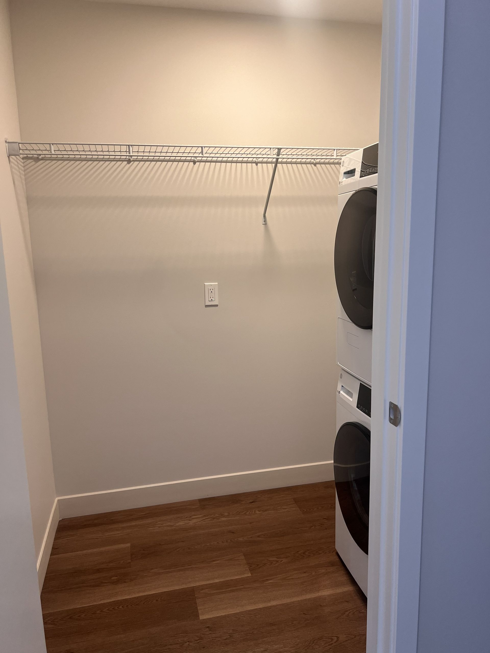 Empty closet with wire shelf, hardwood floor, and stacked washer/dryer on the right.