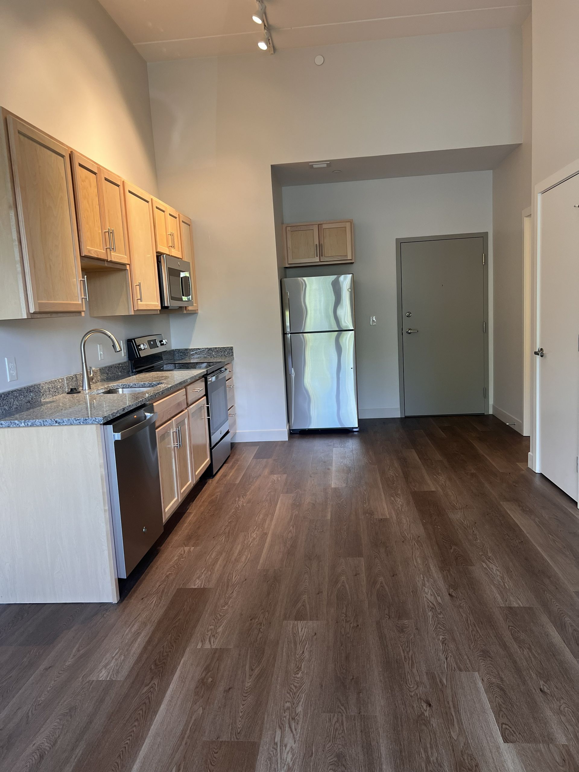 Empty kitchen with wood-look flooring, white cabinets, stainless steel appliances, and a refrigerator in the back.