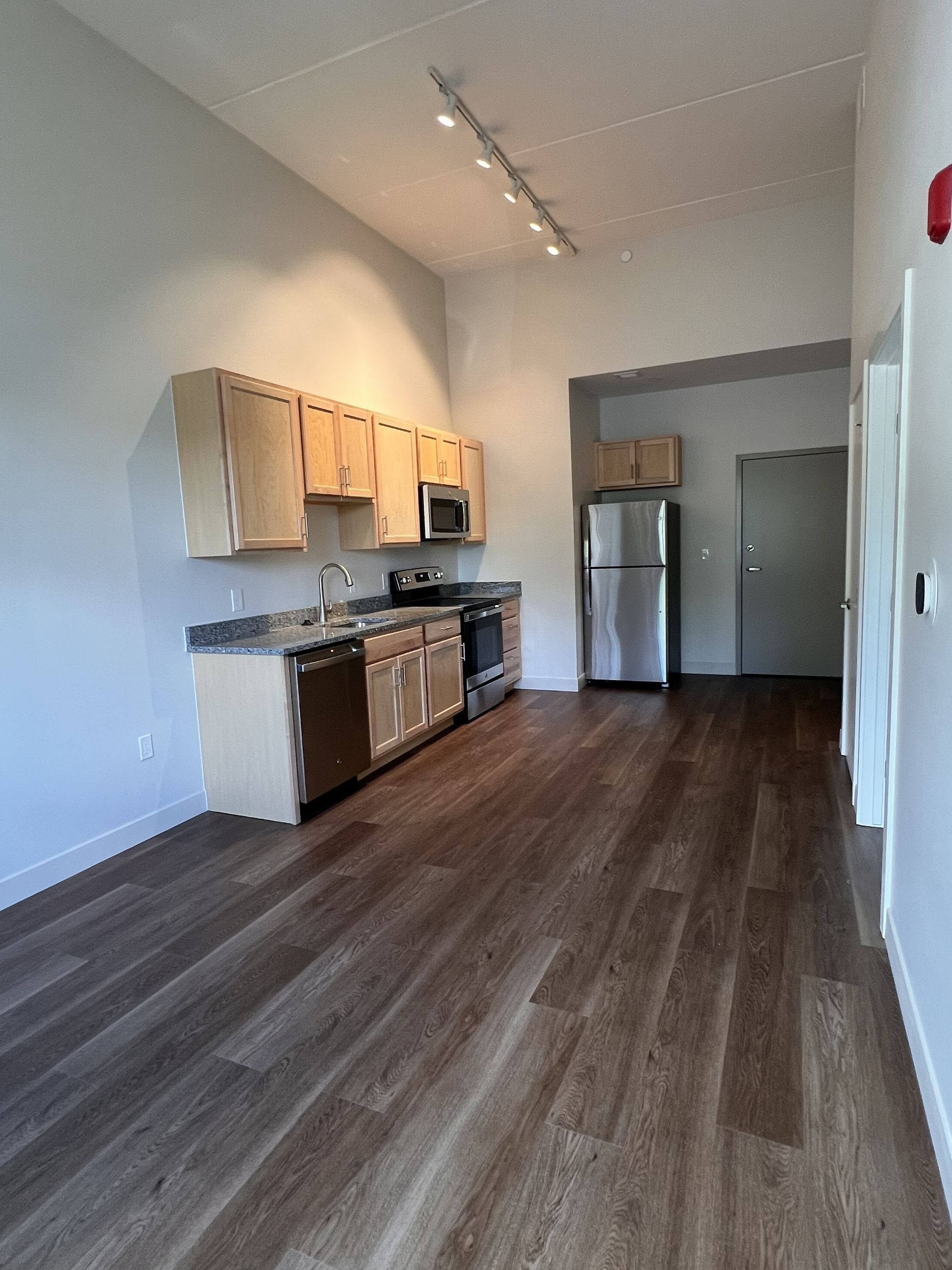 Empty modern kitchen with wood floors, light cabinets, stainless steel appliances, and a refrigerator in the back.
