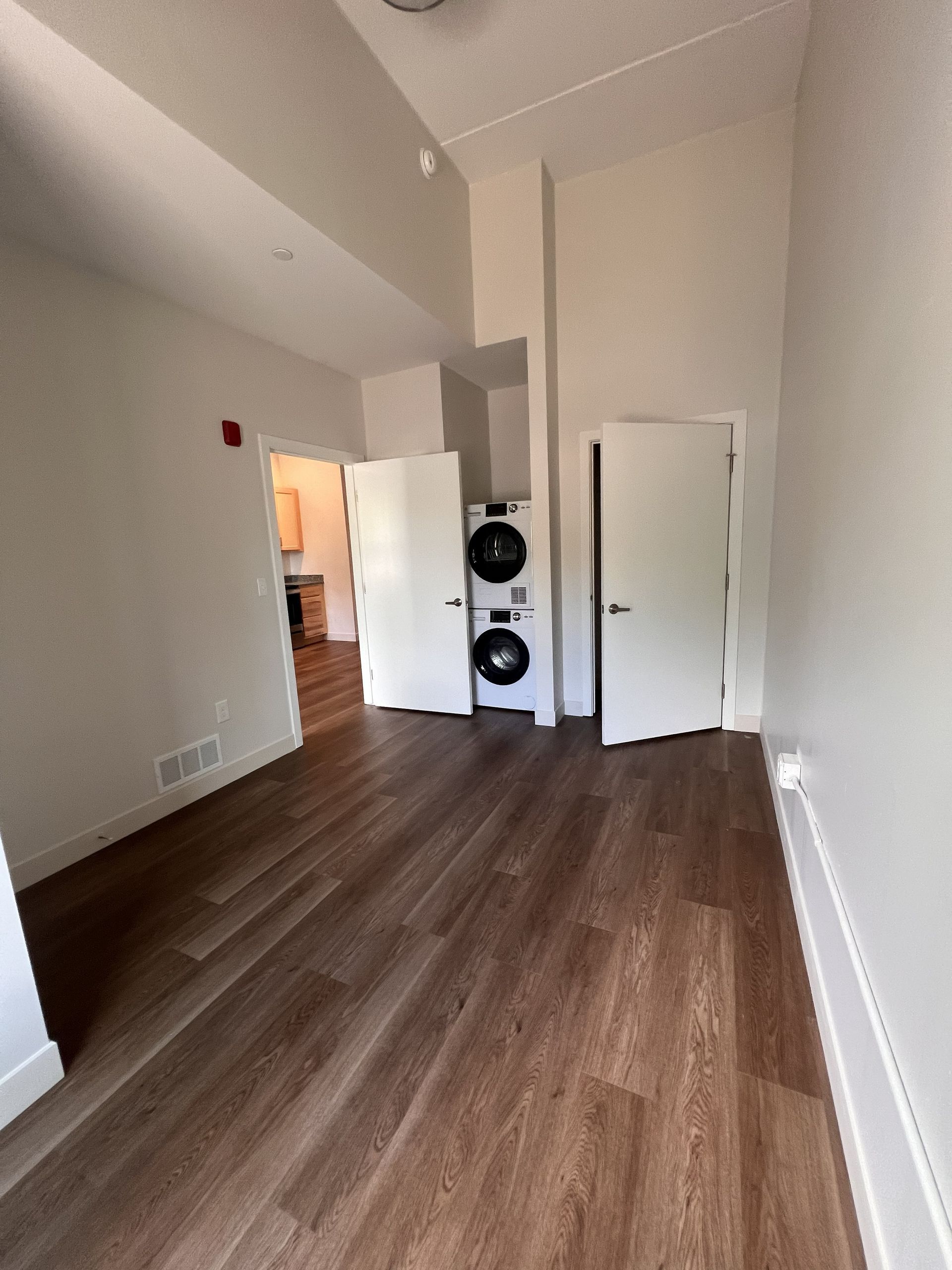 Empty apartment room with wood floors, white walls, washer and dryer, and an open doorway to a lit hallway
