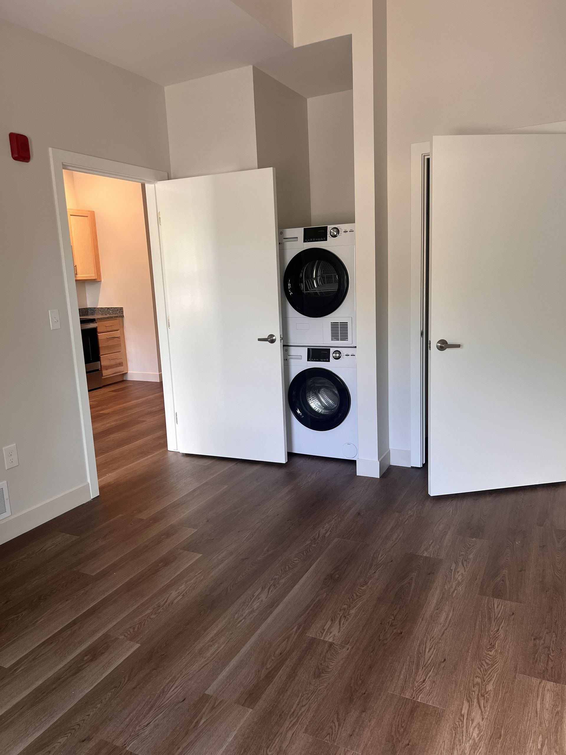 Empty apartment room with hardwood floors, open doors, and stacked washer and dryer in a nook