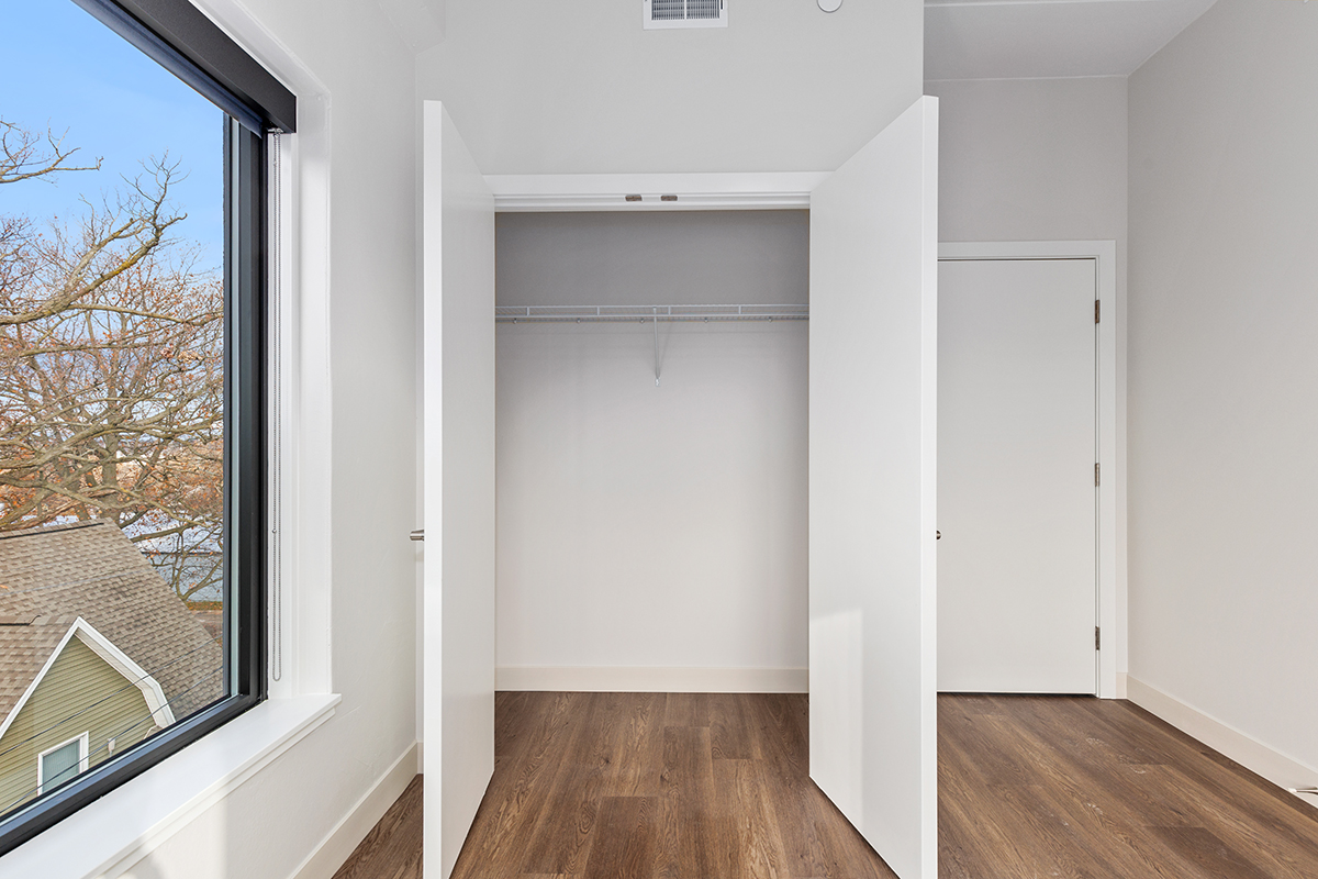 Bright empty bedroom with open closet, white walls, and wood flooring, viewed by a window