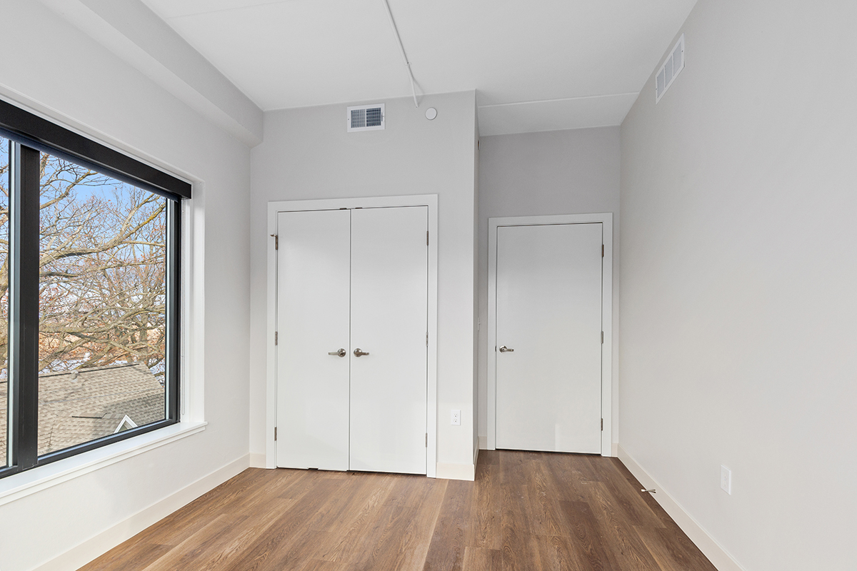 Bright empty bedroom with white walls, wood floor, window, and closet doors