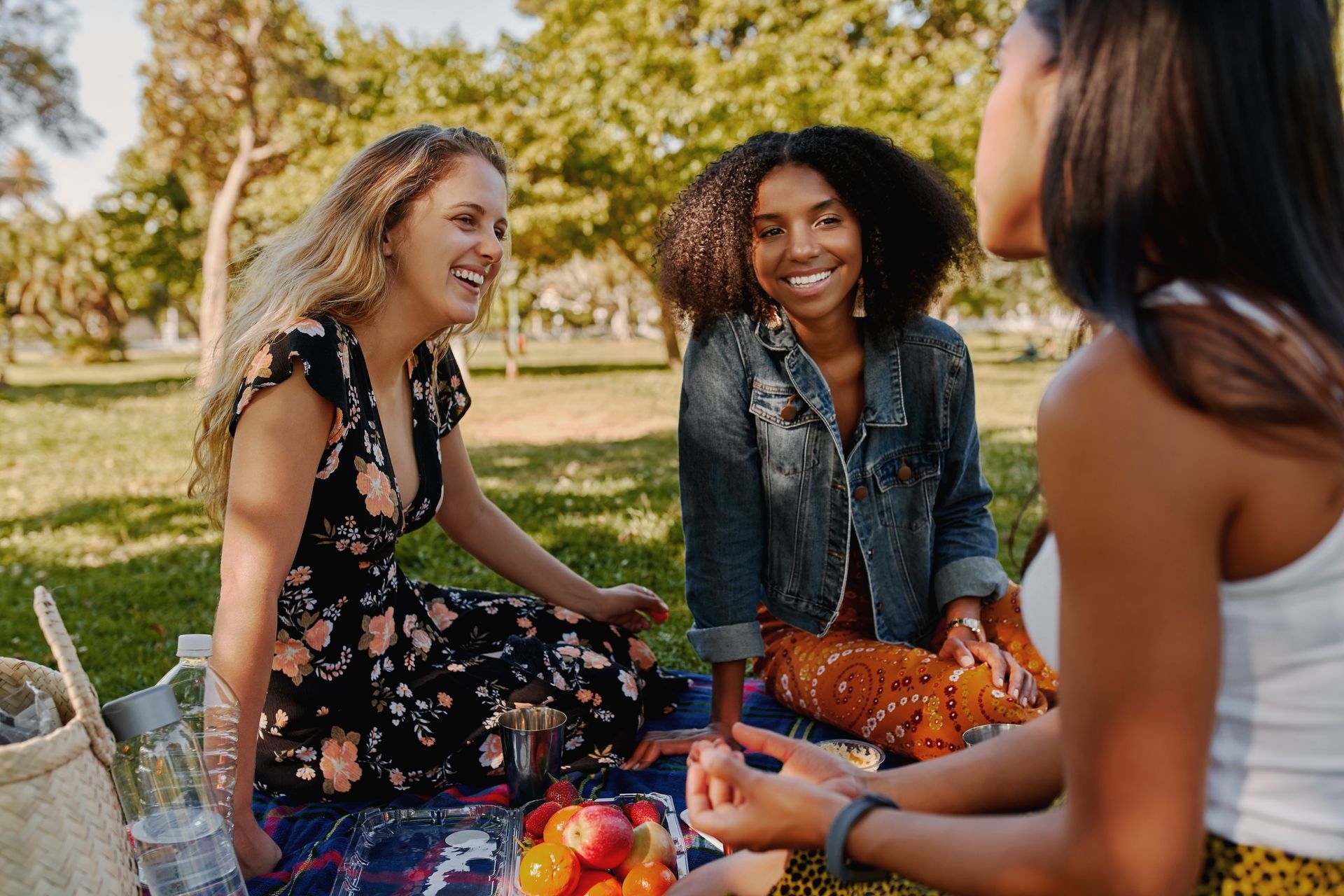 Friends laughing on a picnic blanket in a sunny park, sharing fruit and drinks.