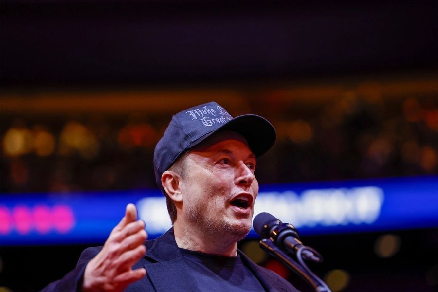 Man speaking into a microphone on stage, wearing a dark cap and shirt, with blurred lights in the background
