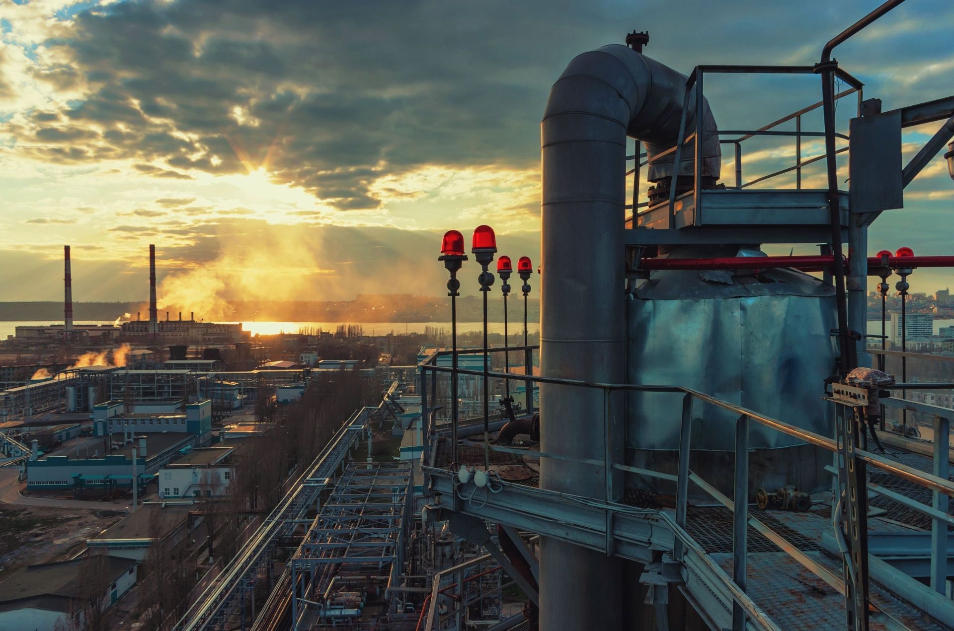 Industrial plant at sunset with pipes, smokestacks, and red warning lights under a glowing sky