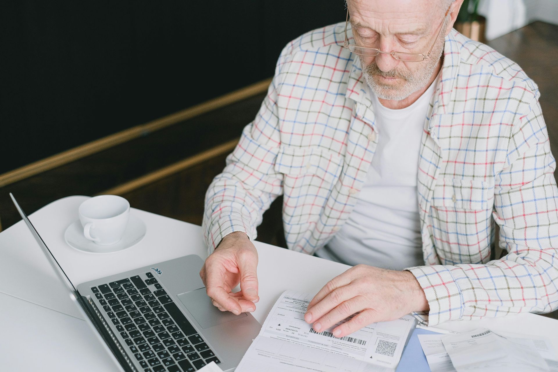 Man working at a desk with a laptop, coffee cup, and papers, reviewing notes with a pen