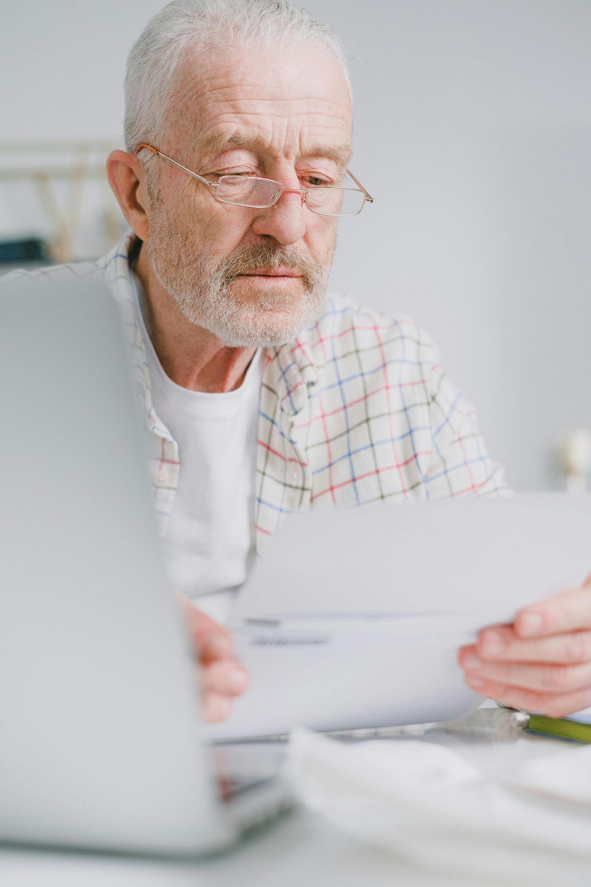 Older man reading a document at a desk beside a laptop, wearing glasses and a plaid shirt