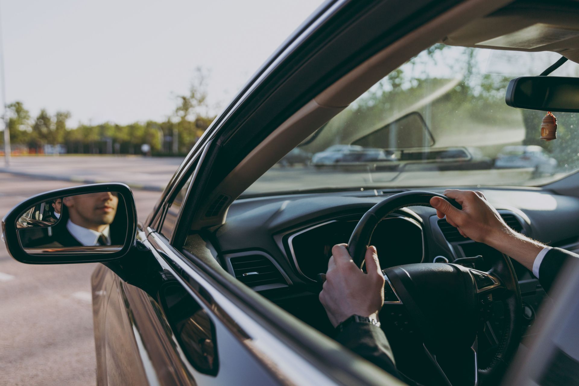 Driver holding steering wheel inside car, seen from side mirror on a sunny road.