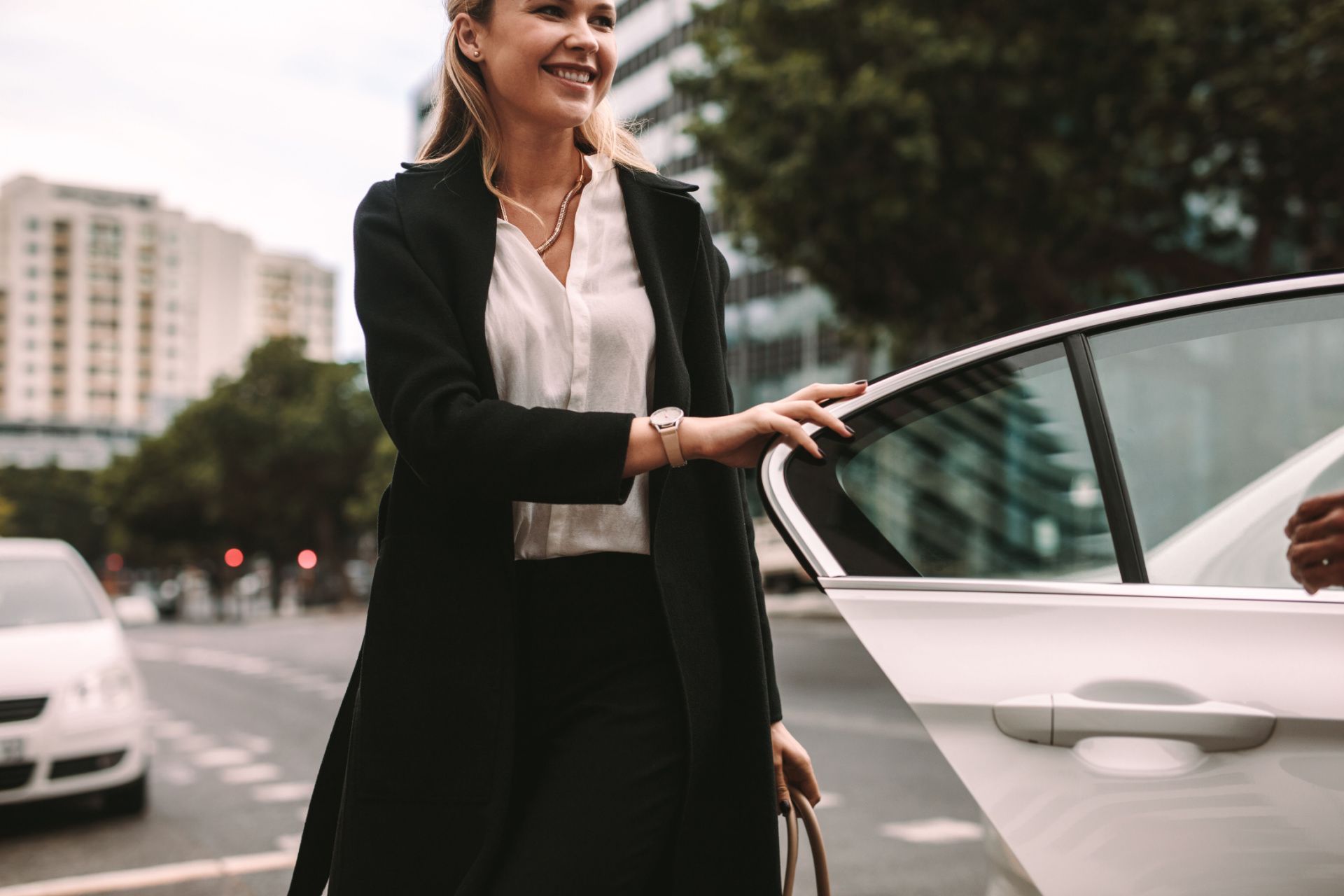 Woman in a black coat smiling beside an open car door on a city street